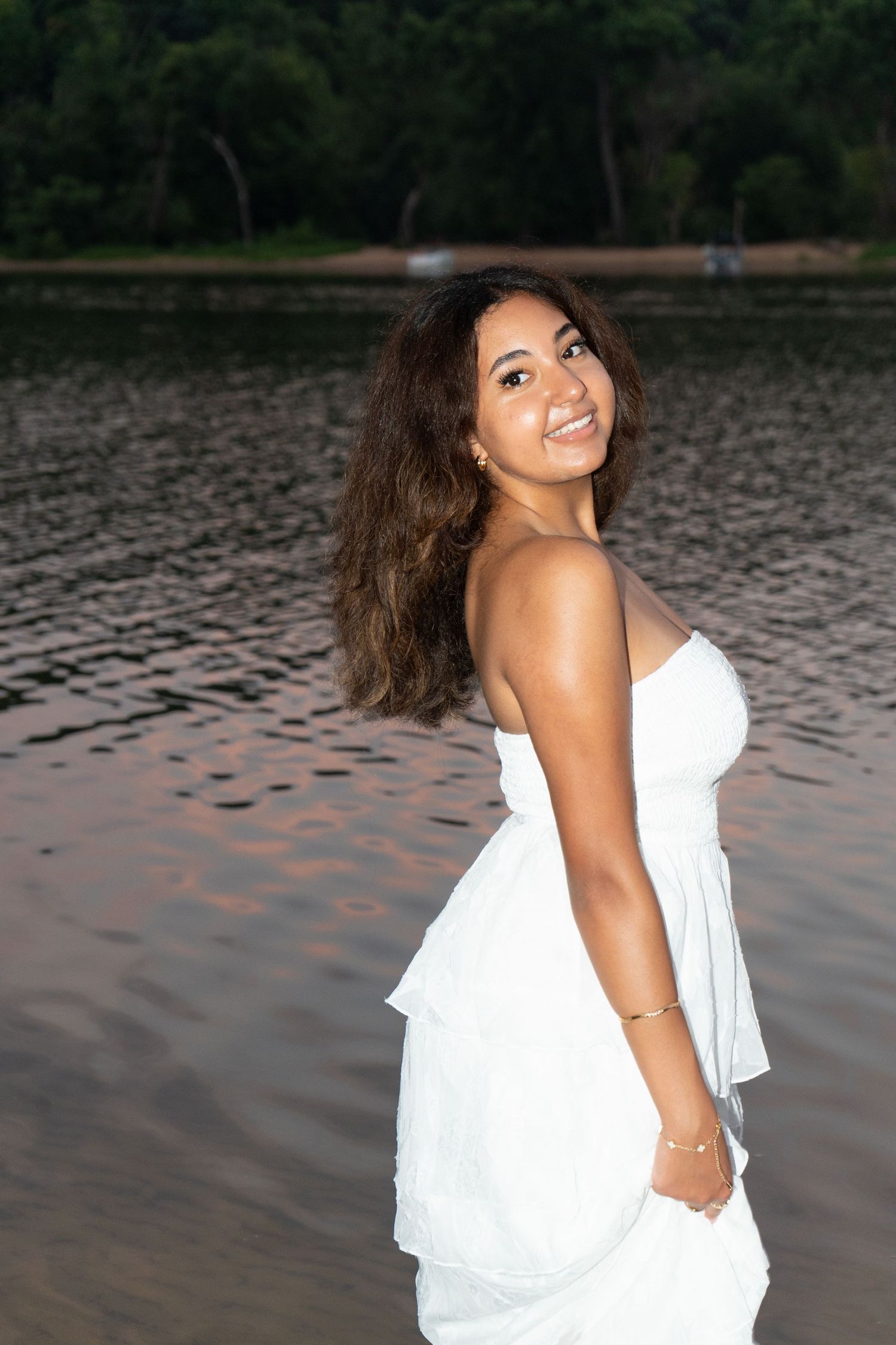 Young woman in white strapless dress smiling by a calm lake with lush green trees in the background.