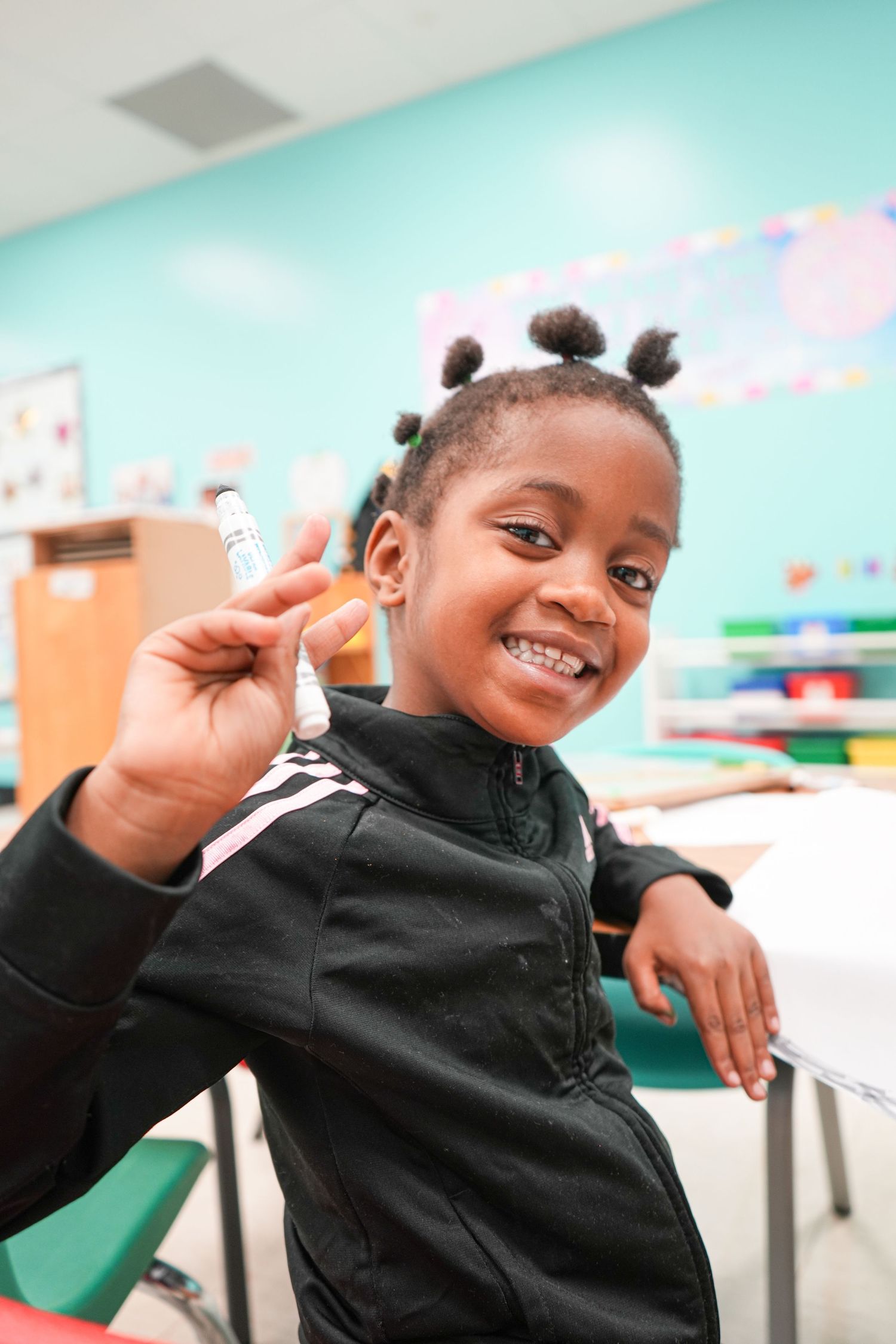 Smiling young girl in a classroom holding a marker, wearing a black jacket, with colorful decorations in the background.
