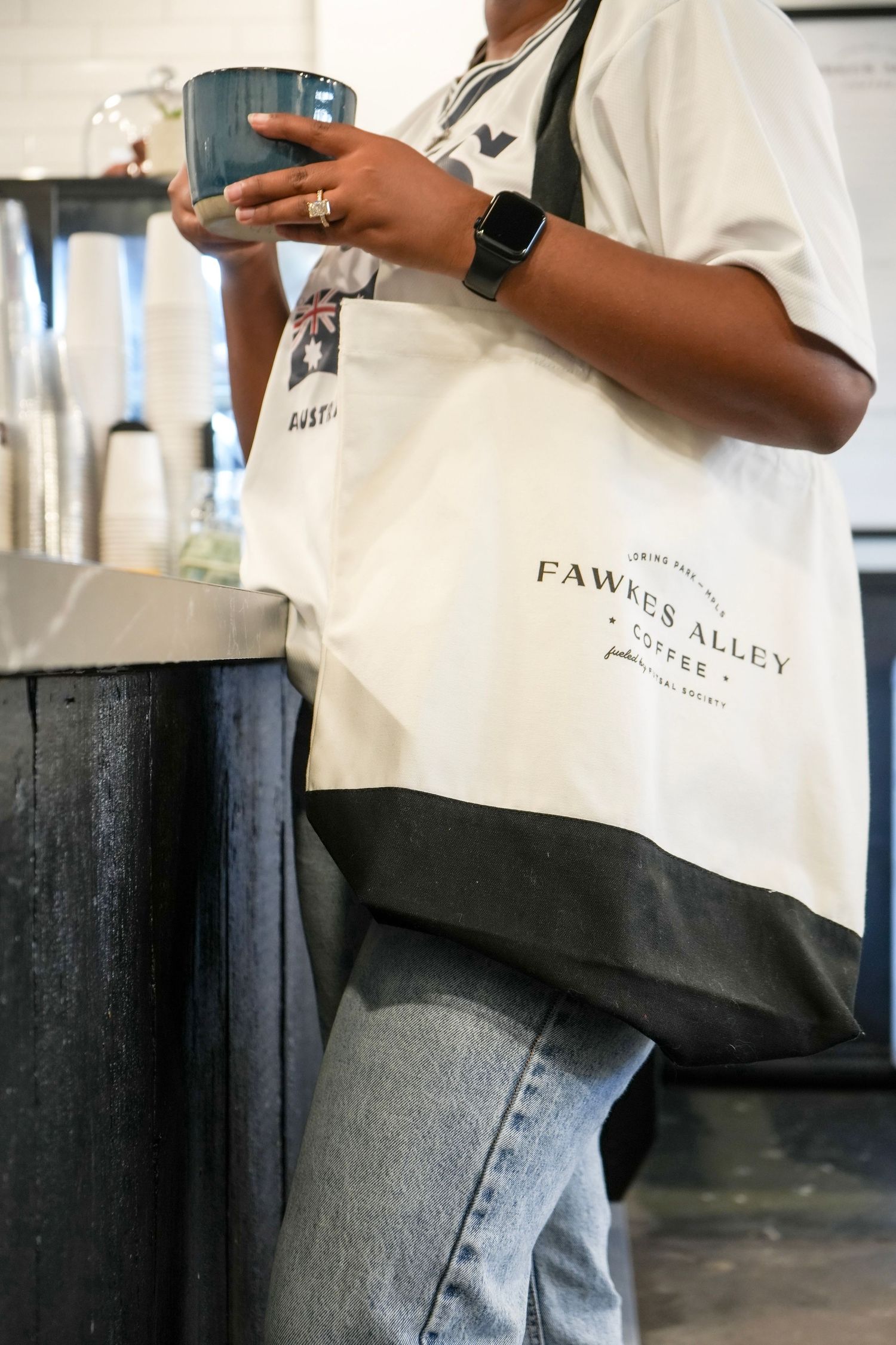 Person holding a mug wearing a Fawkes Alley Coffee canvas tote bag and apple watch in a coffee shop.