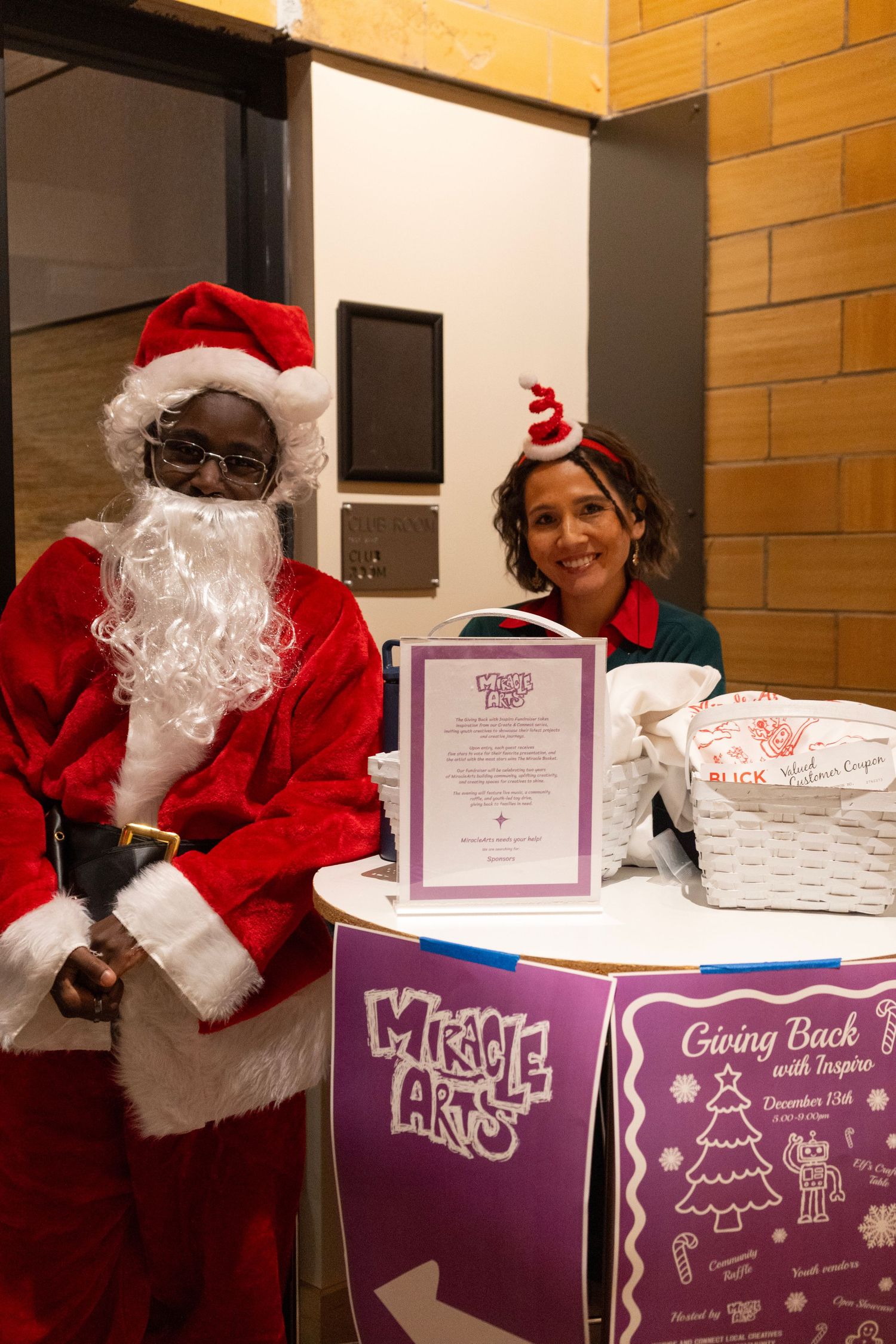 Santa Claus and elf pose at Miracle Arts Giving Back event table with gift baskets on December 15th.