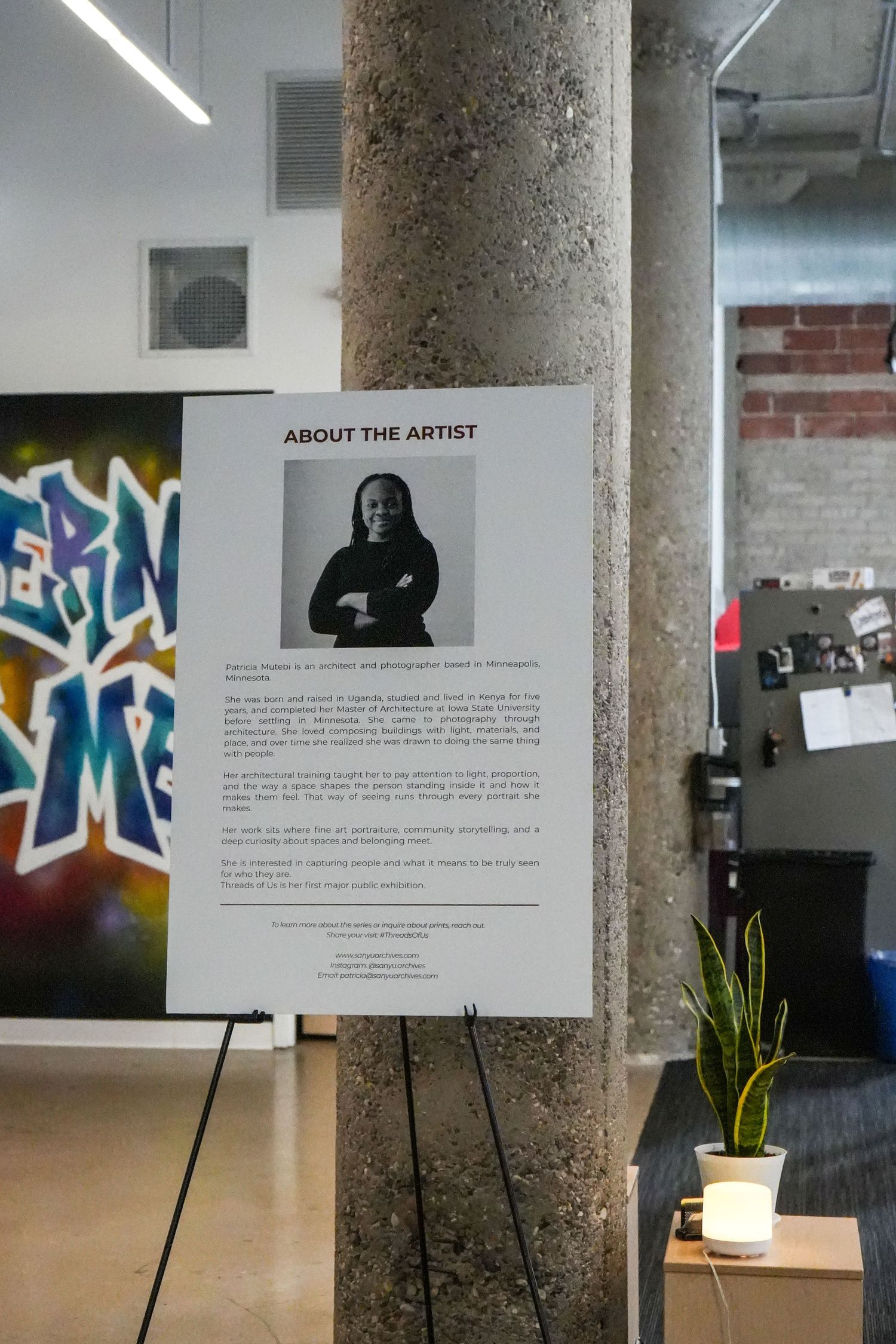 Gallery display of an artist biography poster on an easel beside a graffiti mural in a modern art space.