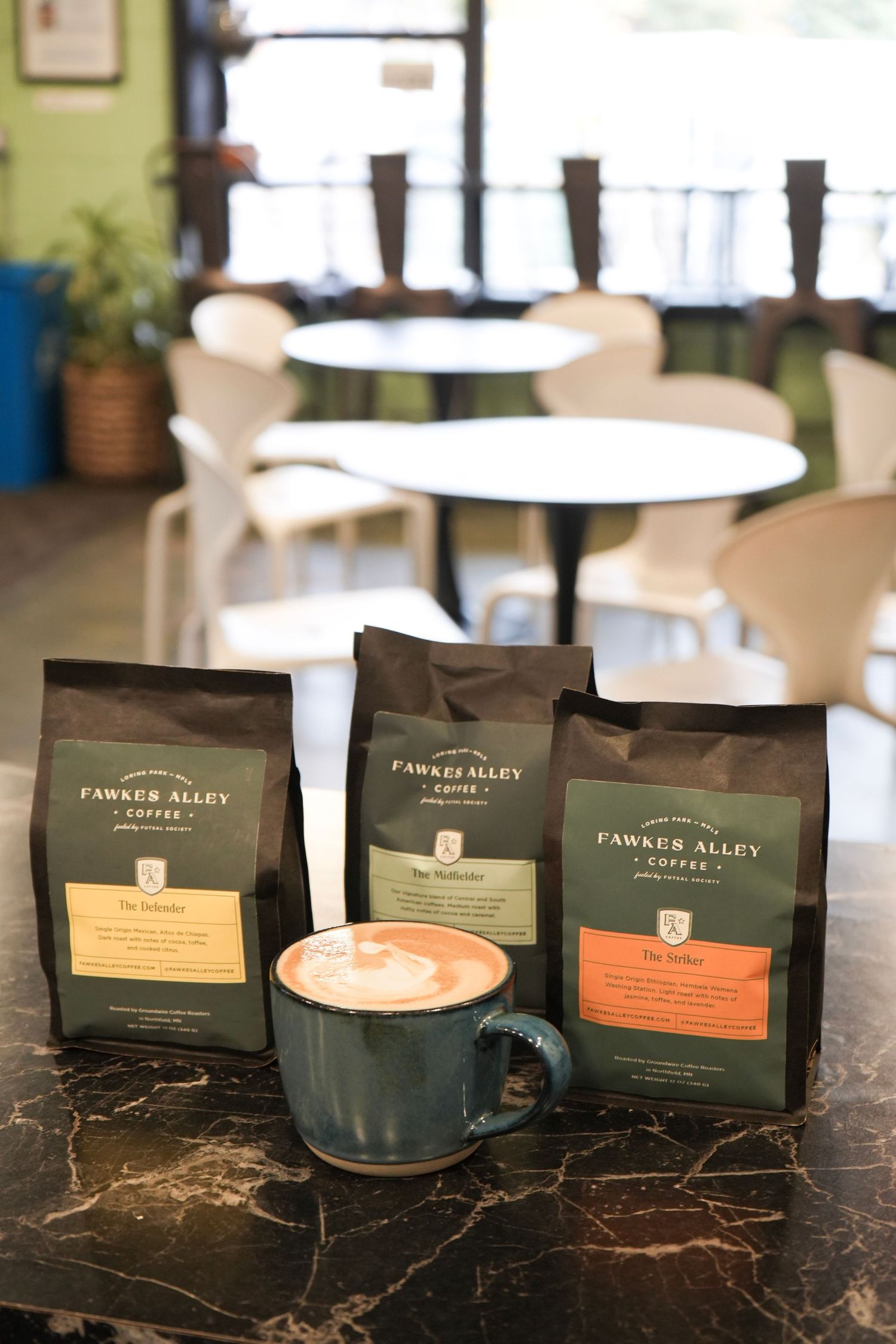 Three Fawkes Alley Coffee bags displayed beside a latte art cappuccino in a blue mug on a marble table in a cafe.