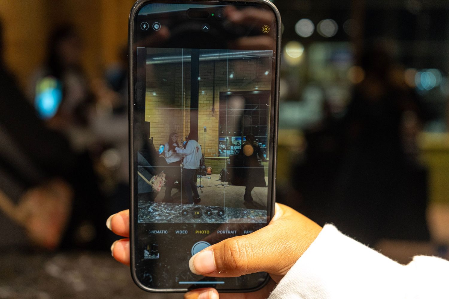 A hand holds a smartphone capturing a photo of people at an elegant indoor event venue through the camera app.