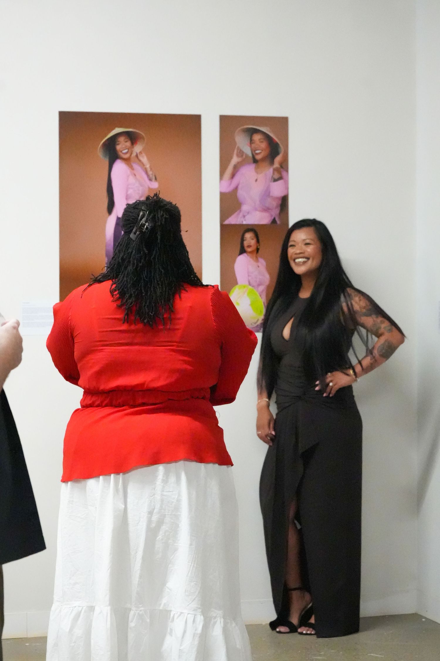 Two women pose near portrait photos at an art gallery, one in red dress, one in black jumpsuit with tattoos.