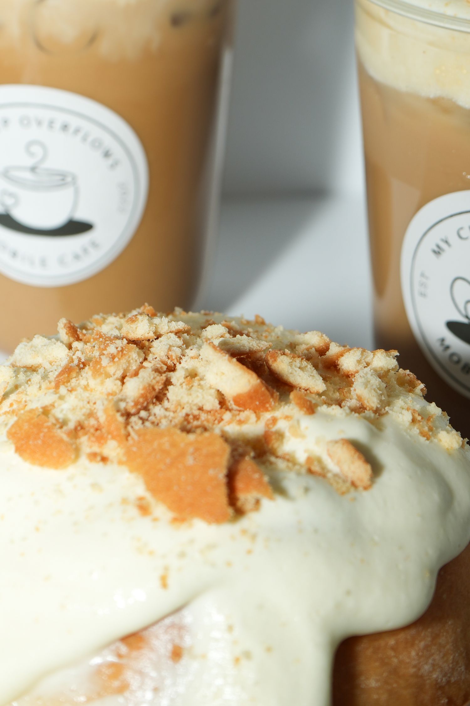 Close-up of a creamy dessert topped with crumbles and iced coffee drinks from My Overflow Cafe in background.