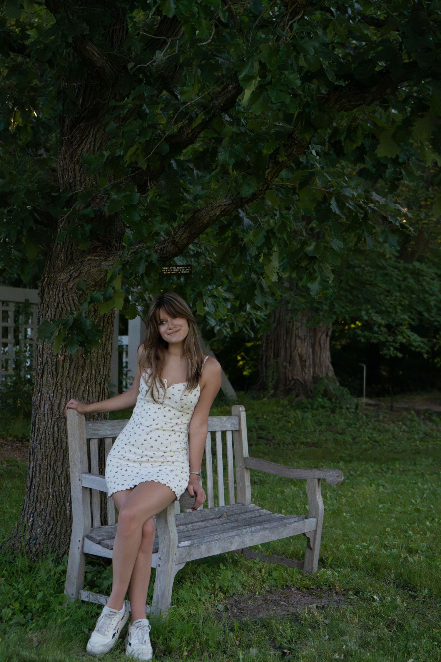 Young woman in white floral sundress sitting on wooden garden bench under large green tree.