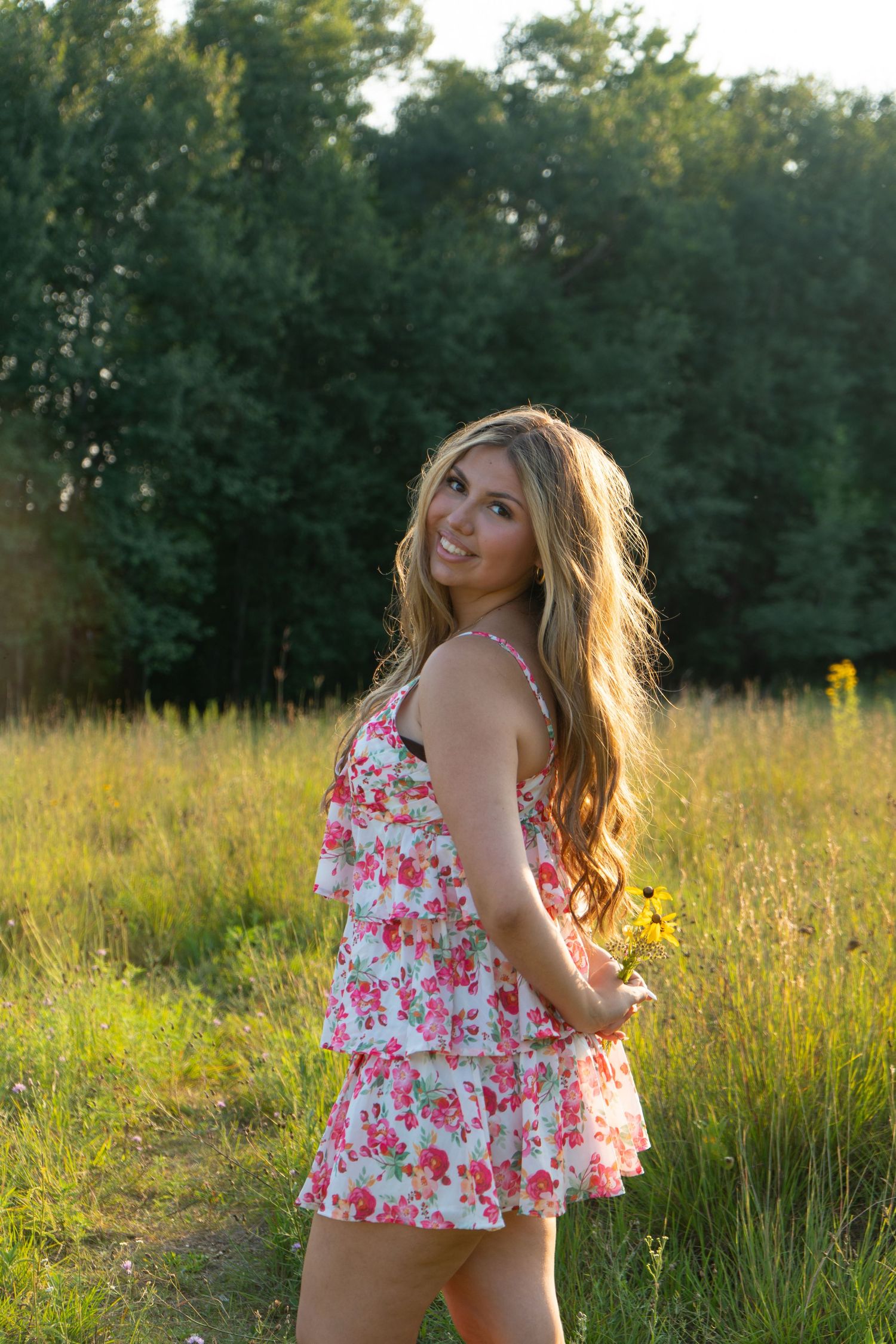Young woman in floral sundress holding wildflowers, smiling in a sunny green meadow with trees in background.