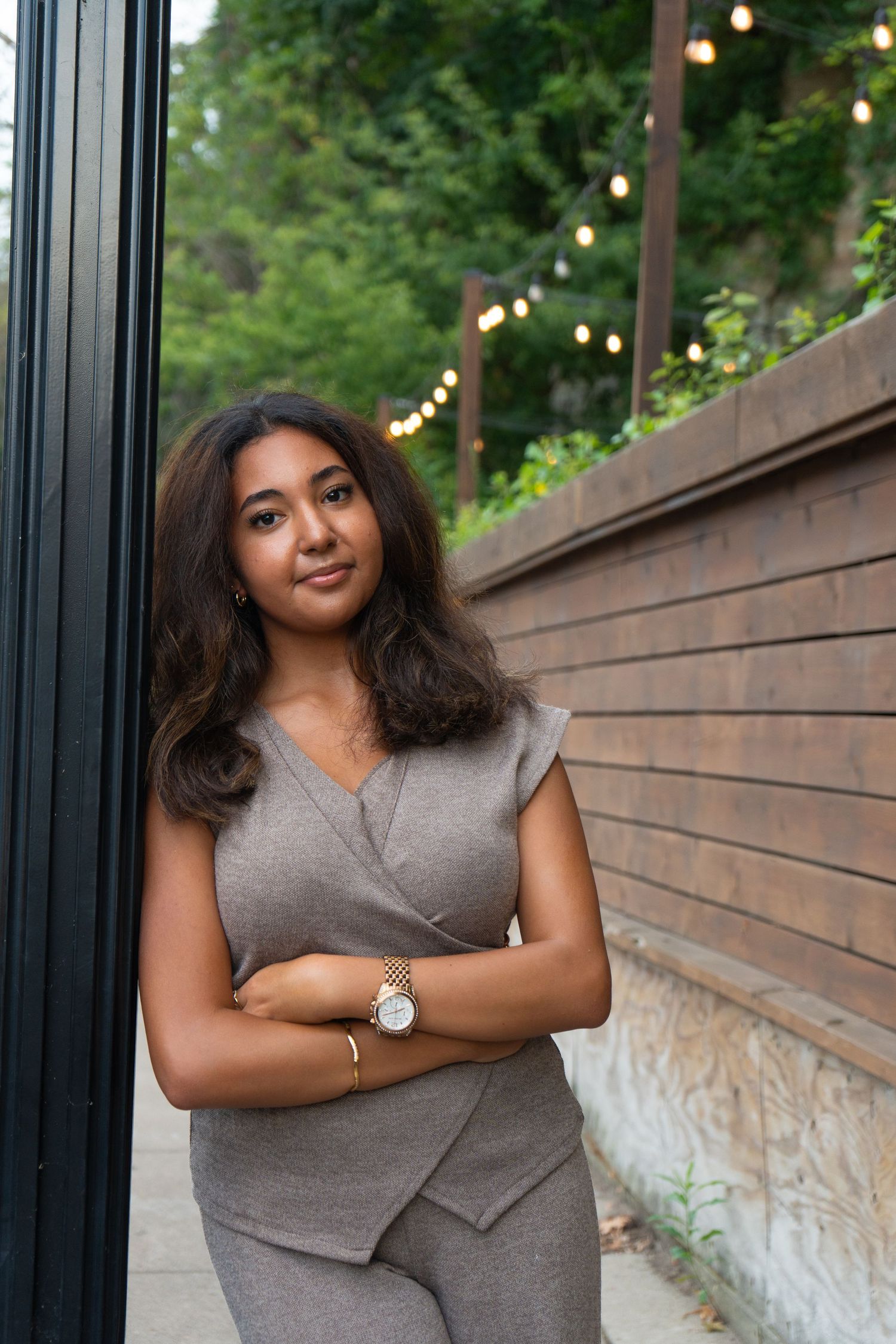 A young woman in a gray wrap jumpsuit with arms crossed, posing outdoors near string lights and wooden wall.
