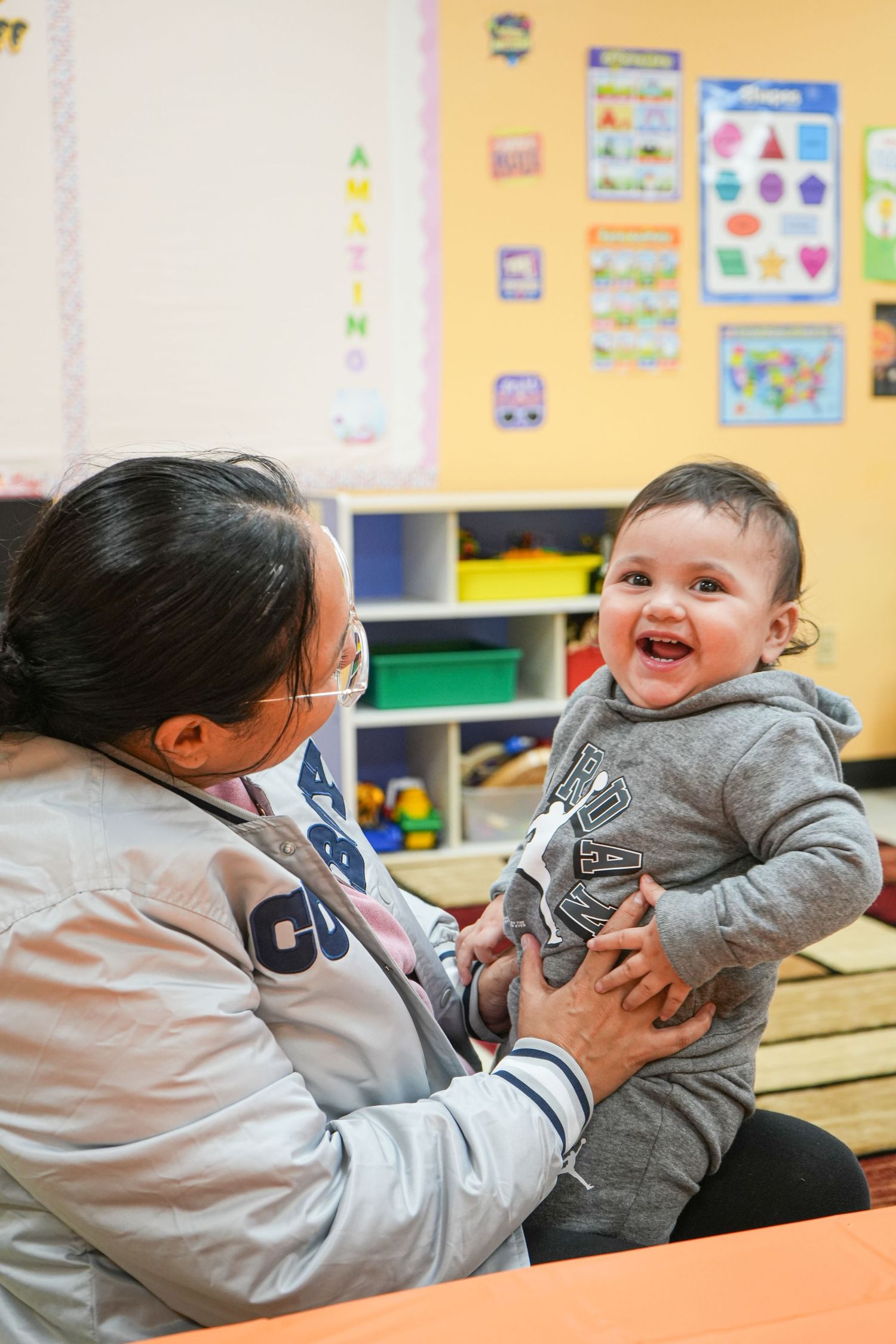 A caregiver holds a smiling baby in a colorful classroom setting, sharing a joyful moment together.