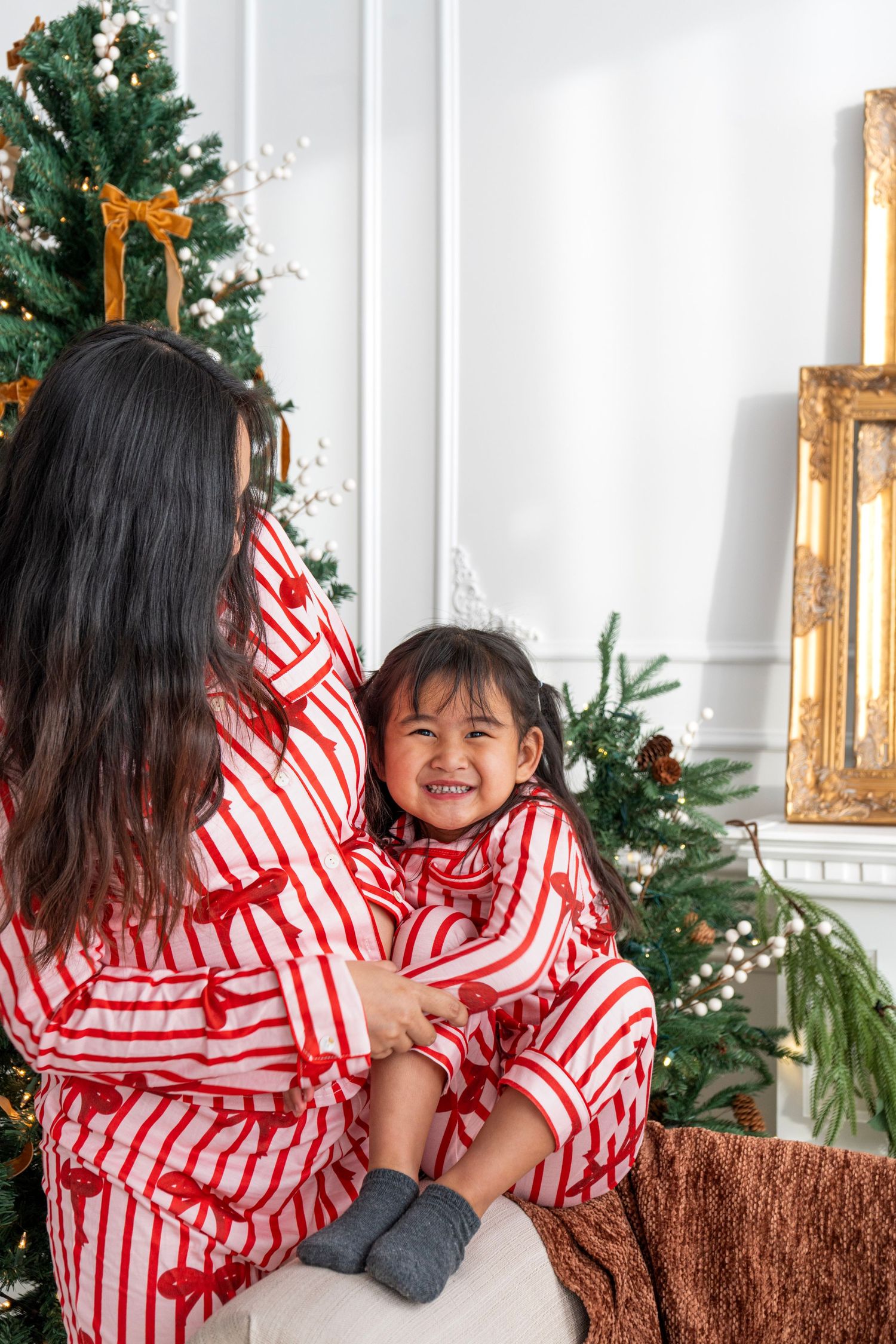 Mother and daughter in matching red striped Christmas pajamas smiling together near a decorated holiday tree.