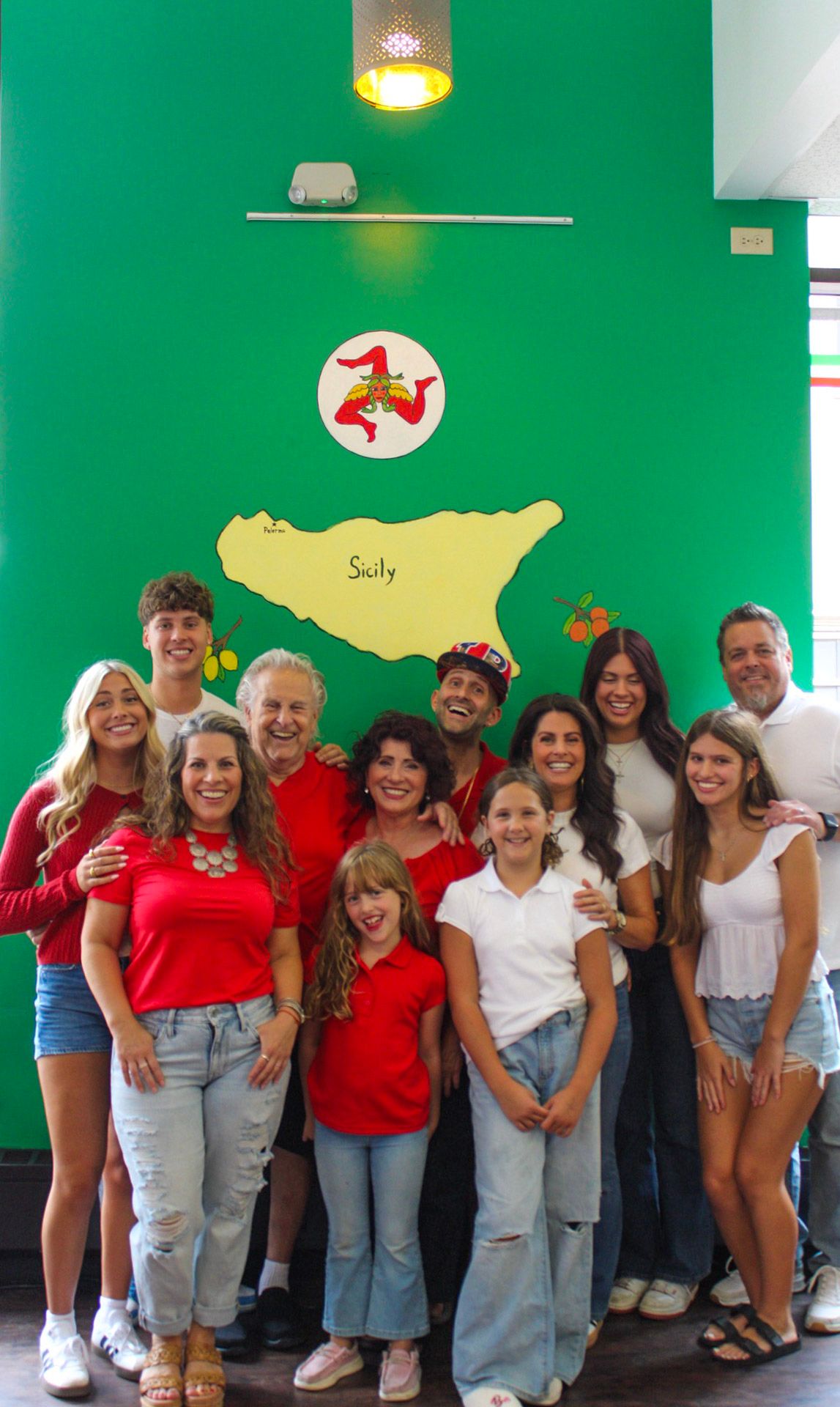 Large family group smiling together in front of a green wall with a Sicily map mural at an Italian restaurant.