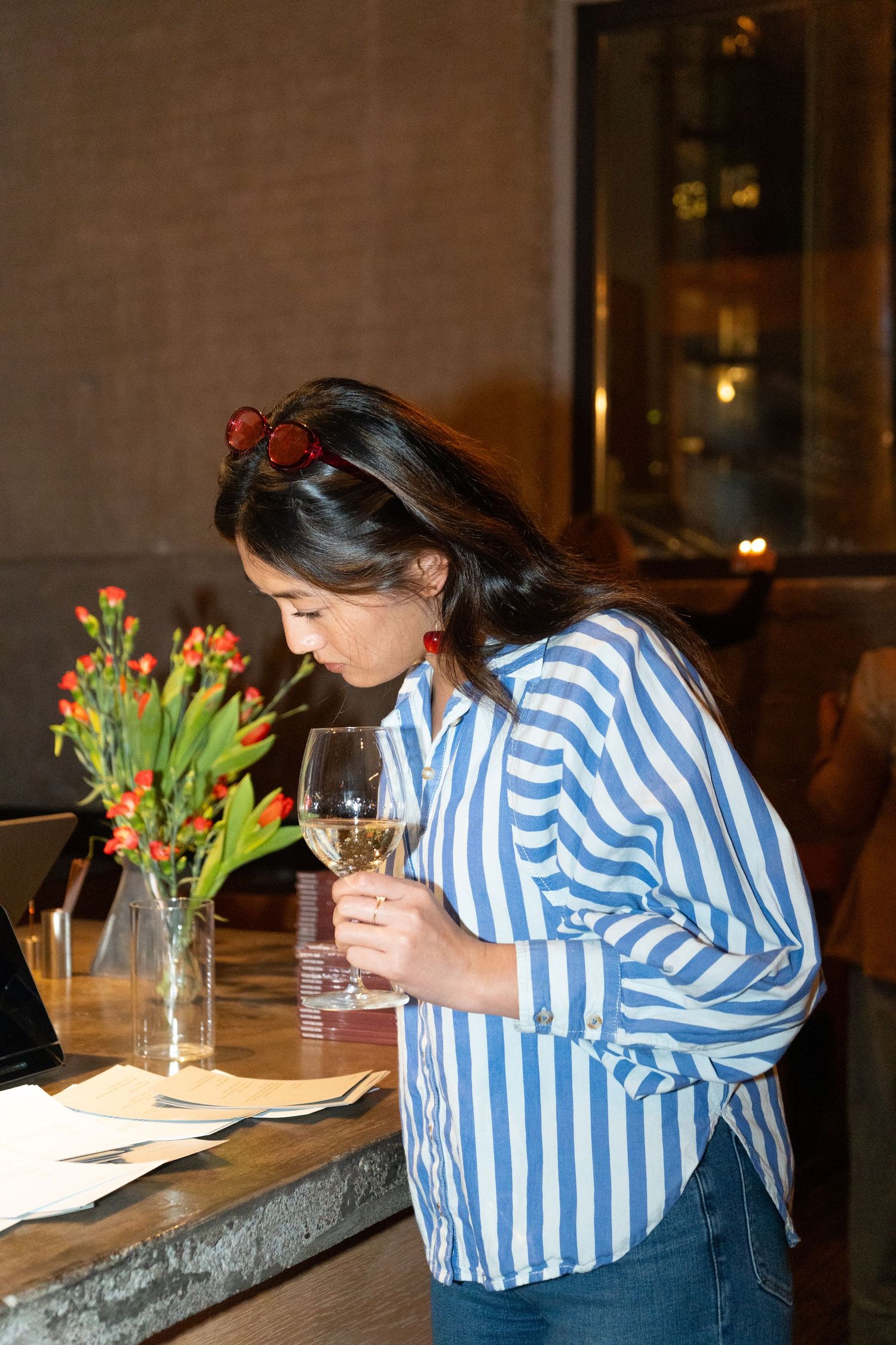 Woman in striped shirt smelling white wine at restaurant table with red tulips.