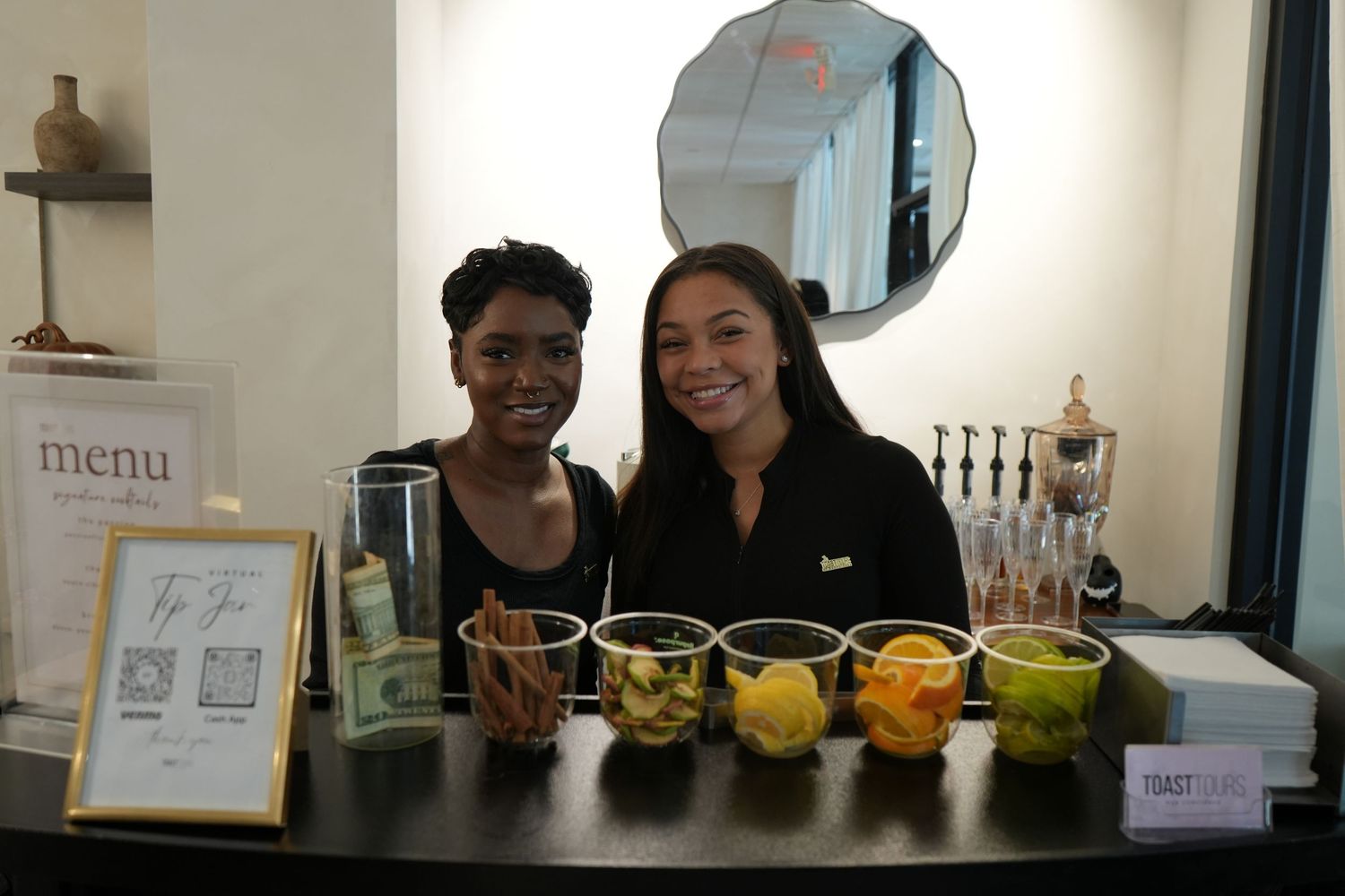 Two smiling women stand behind a bar counter with fresh fruit garnishes and a menu sign at an elegant event.