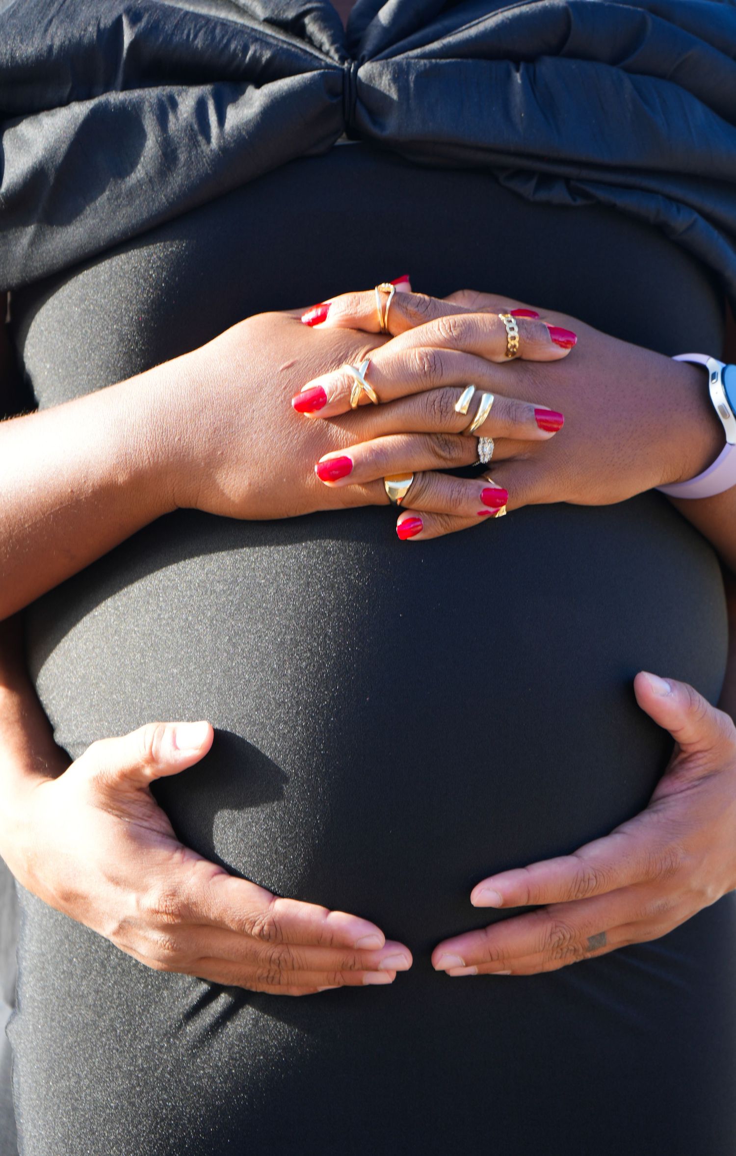 Pregnant woman in black outfit with partner's hands cradling baby bump, red manicured nails and gold rings visible.