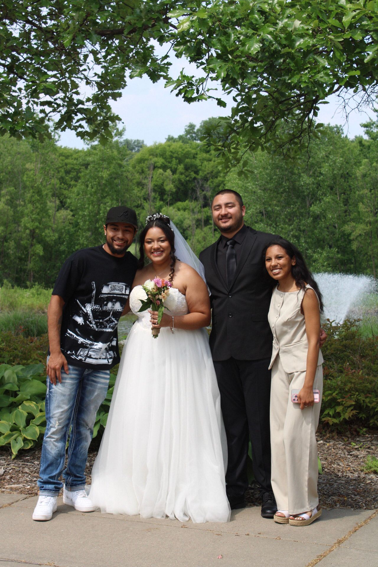 Wedding couple poses with two guests outdoors near a fountain, bride in white gown holding bouquet.