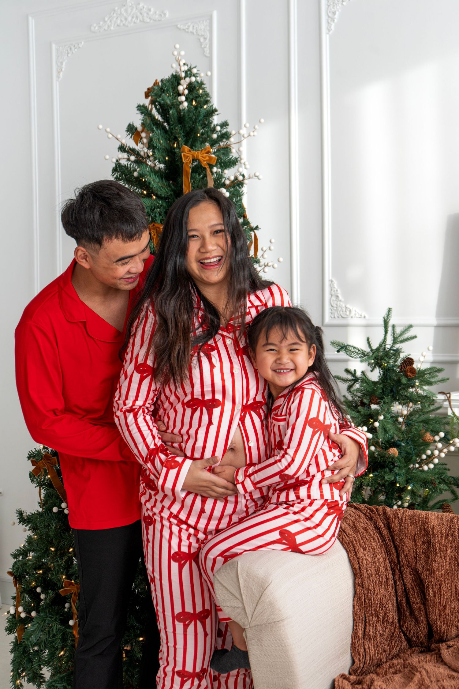 Asian family in matching Christmas pajamas laughing together in front of decorated Christmas trees.