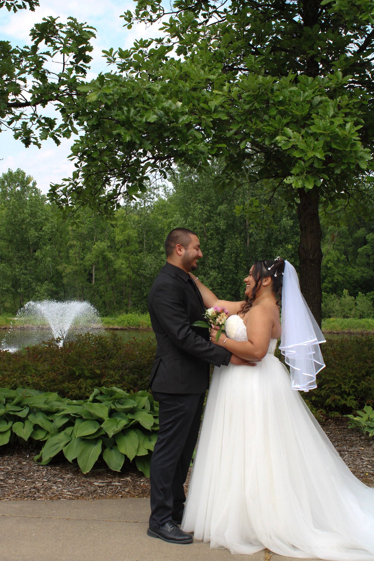 Bride and groom share a romantic moment in a garden park with a fountain and lush green trees in the background.