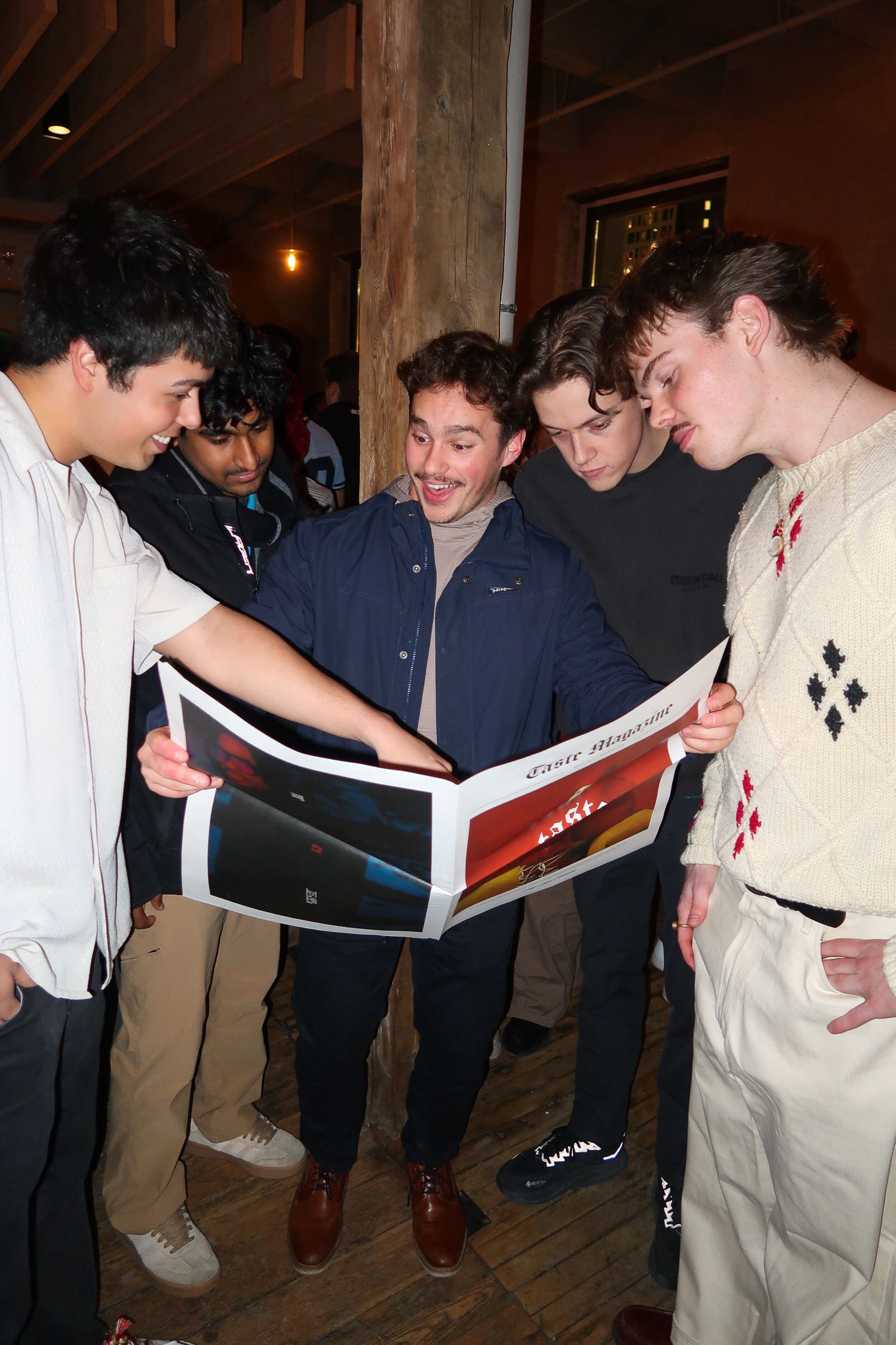 Group of young men gathered together reviewing a large printed photograph or magazine at an indoor event.