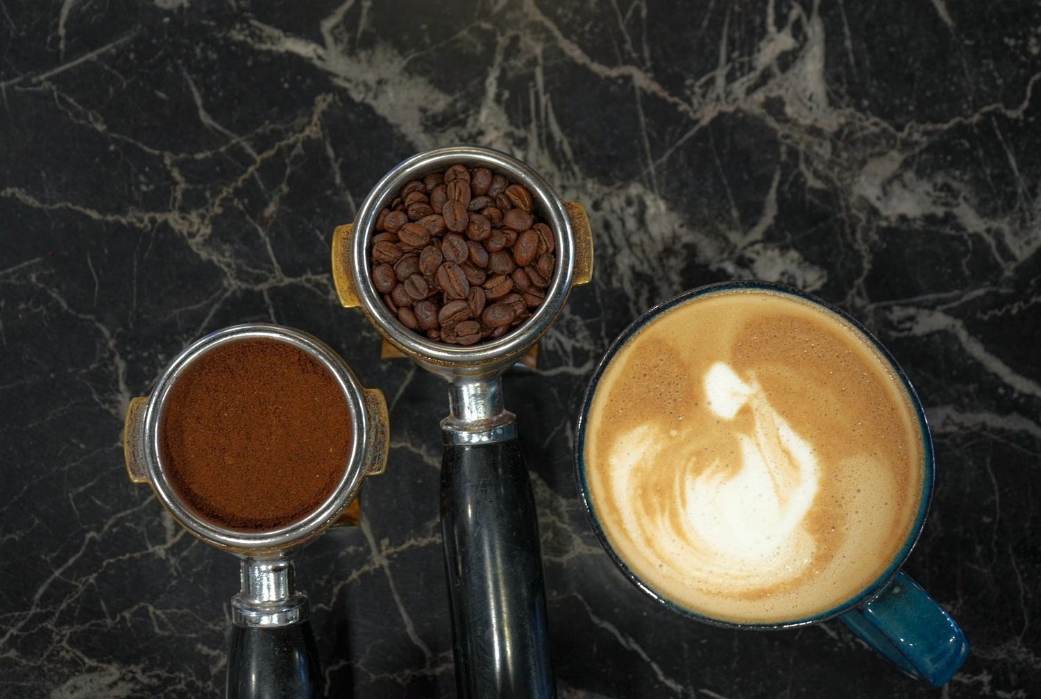 Two espresso portafilters with coffee grounds and beans beside a frothy latte art cappuccino on marble surface.