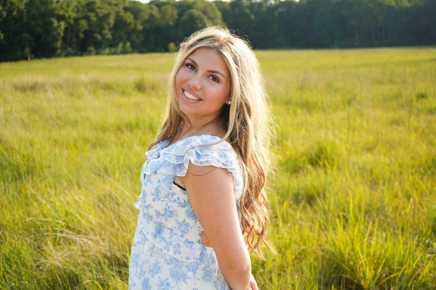 Smiling young woman in a blue floral dress standing in a lush green field on a sunny day.