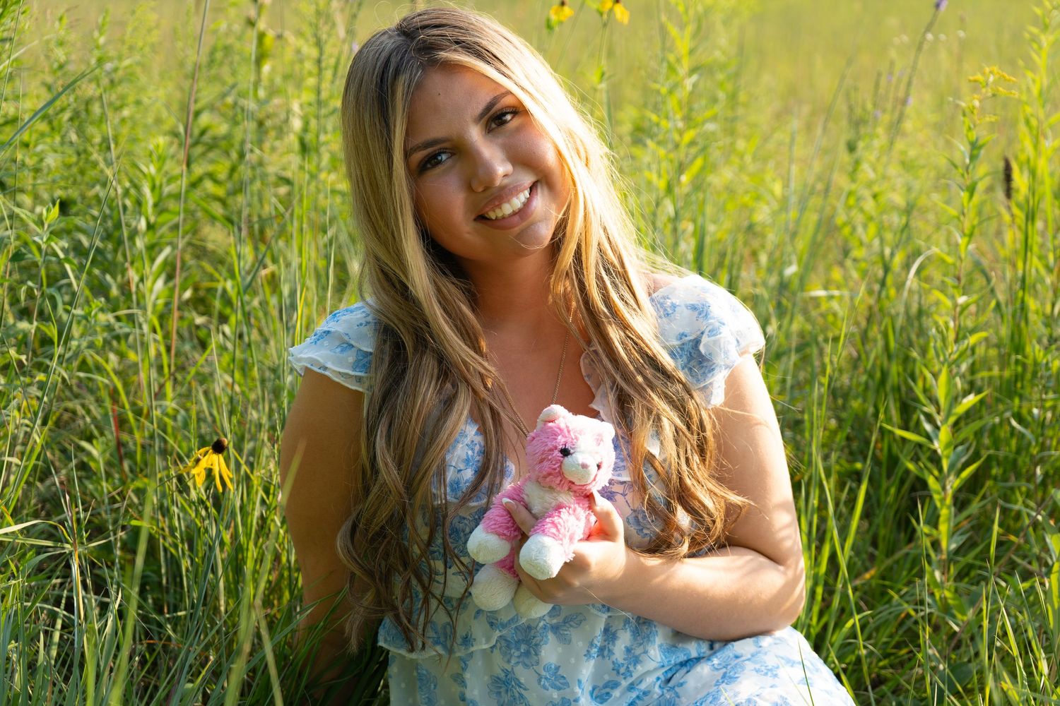Smiling young woman with long blonde hair holding a pink teddy bear while sitting in a sunny green field.