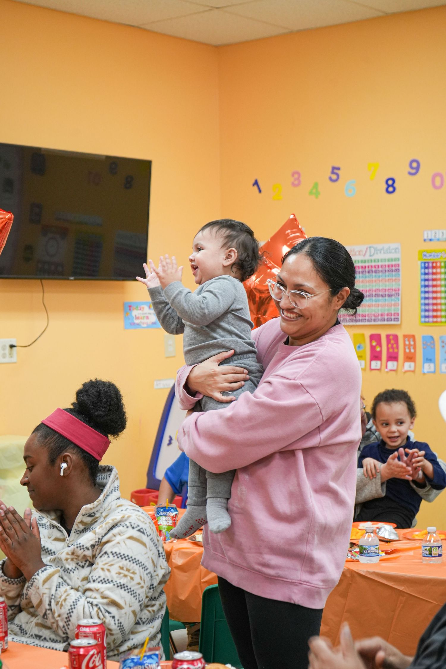 Woman in pink sweatshirt holds smiling baby at colorful children's birthday party with orange decorations.