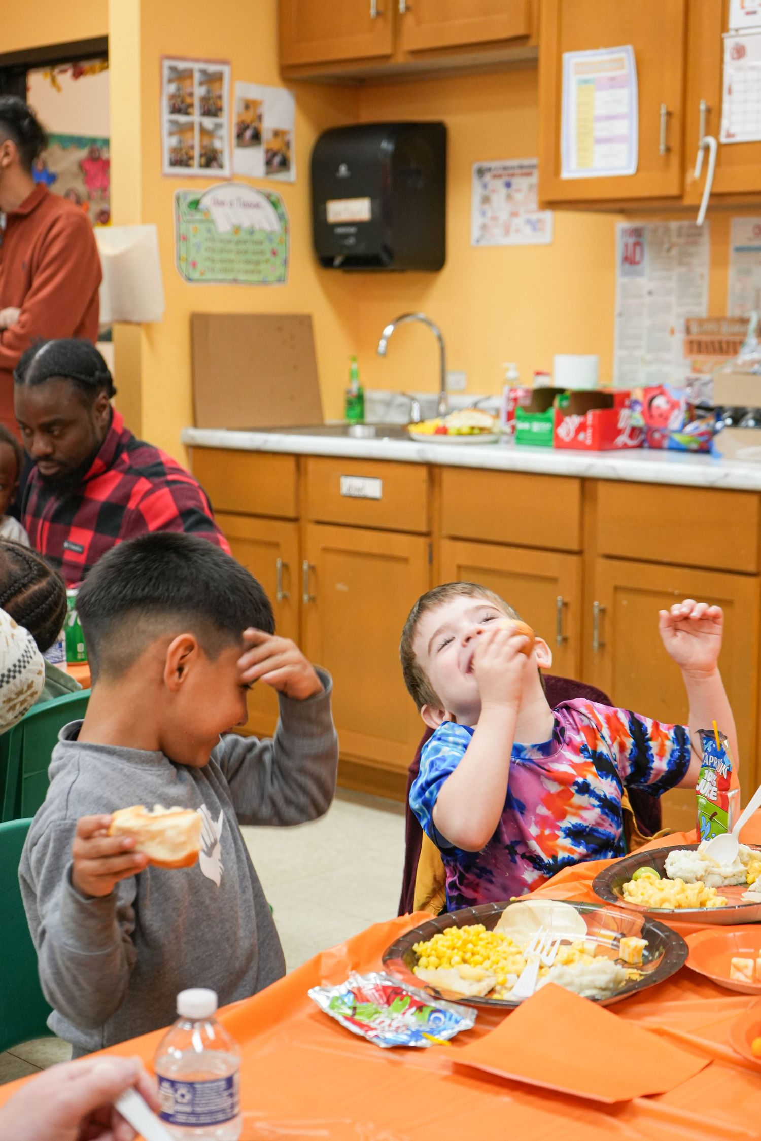 Two children eating at an orange table in a classroom kitchen, one joyfully tossing food in the air.