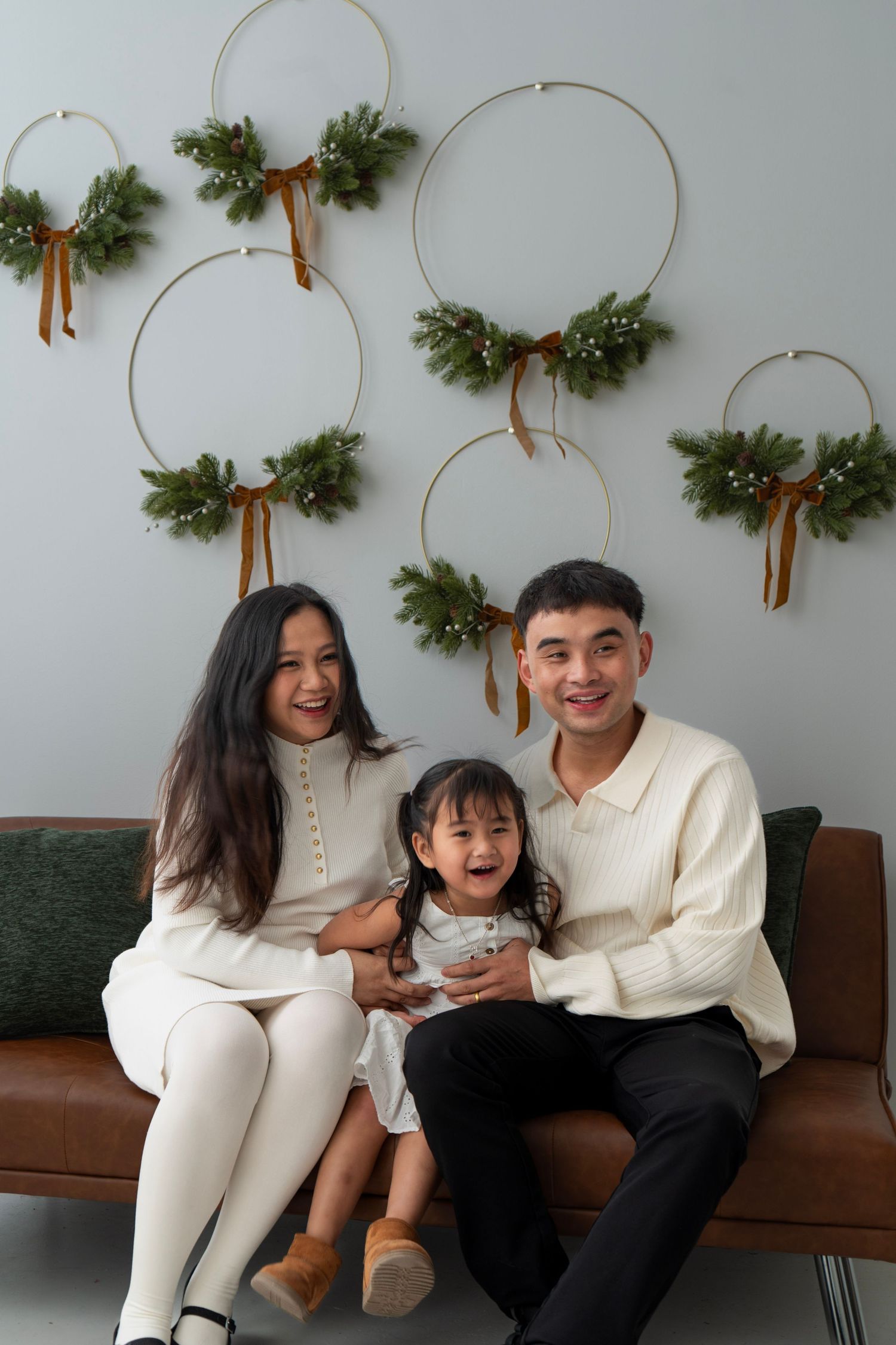 Happy Asian family of three sitting on leather sofa with Christmas wreath wall decor, wearing white outfits for holiday portrait.
