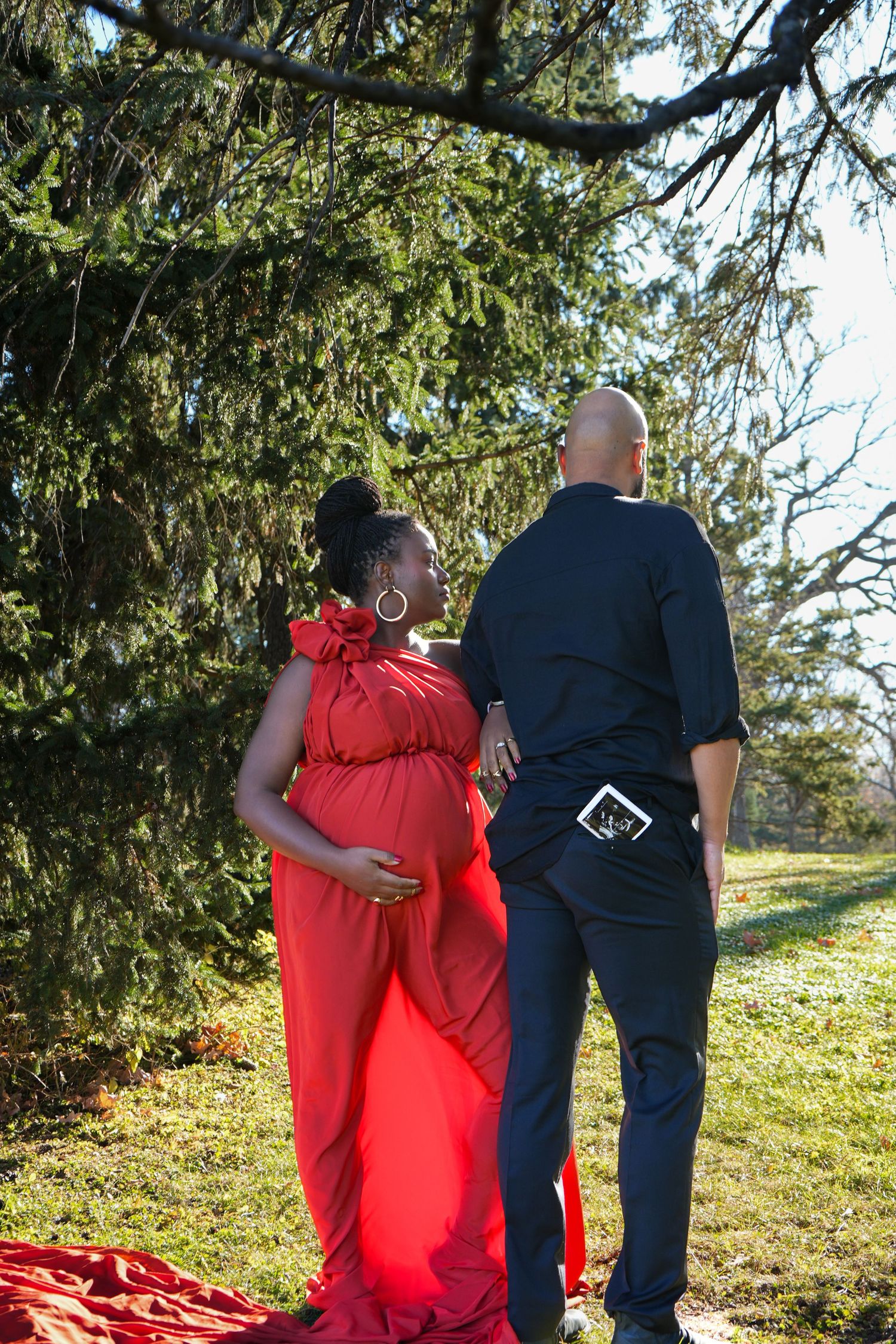 Pregnant woman in red gown with partner standing under trees in outdoor maternity photo shoot.