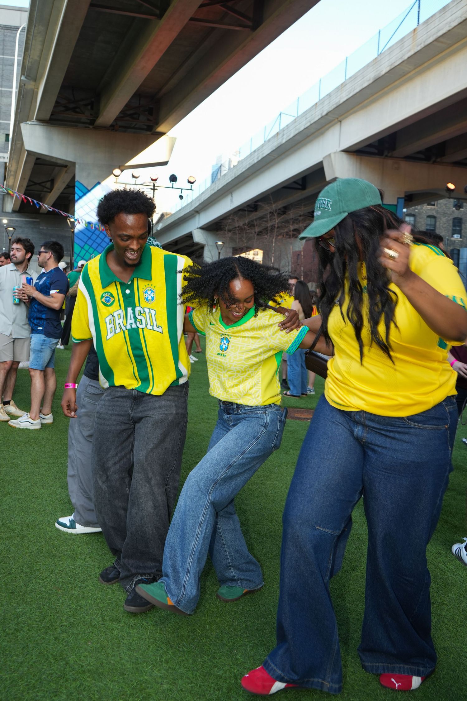 Three people wearing Brazil soccer jerseys dancing outdoors under an overpass on artificial turf.
