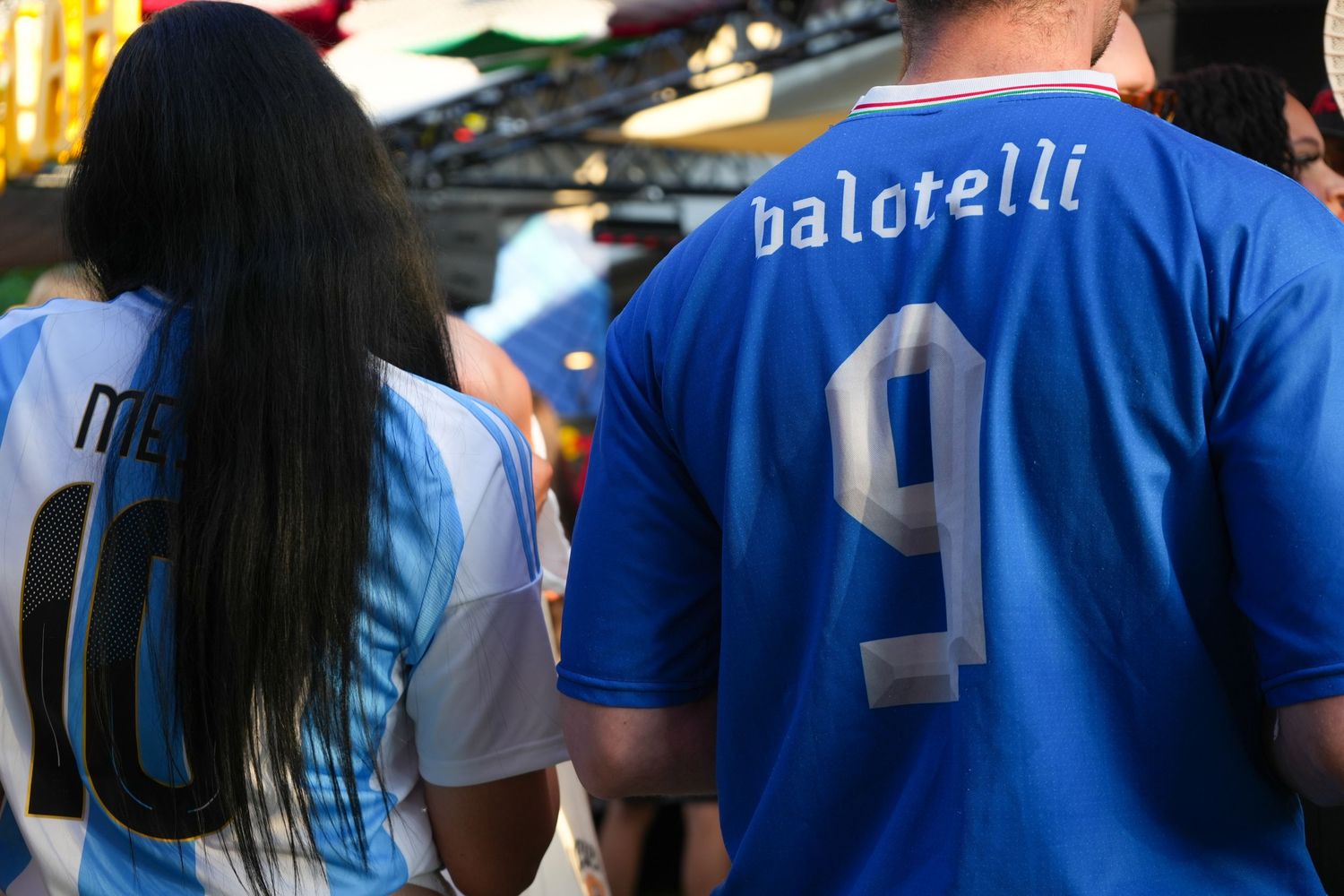 Two soccer fans wearing Messi Argentina and Balotelli Italy number 9 jerseys at a crowded outdoor event.