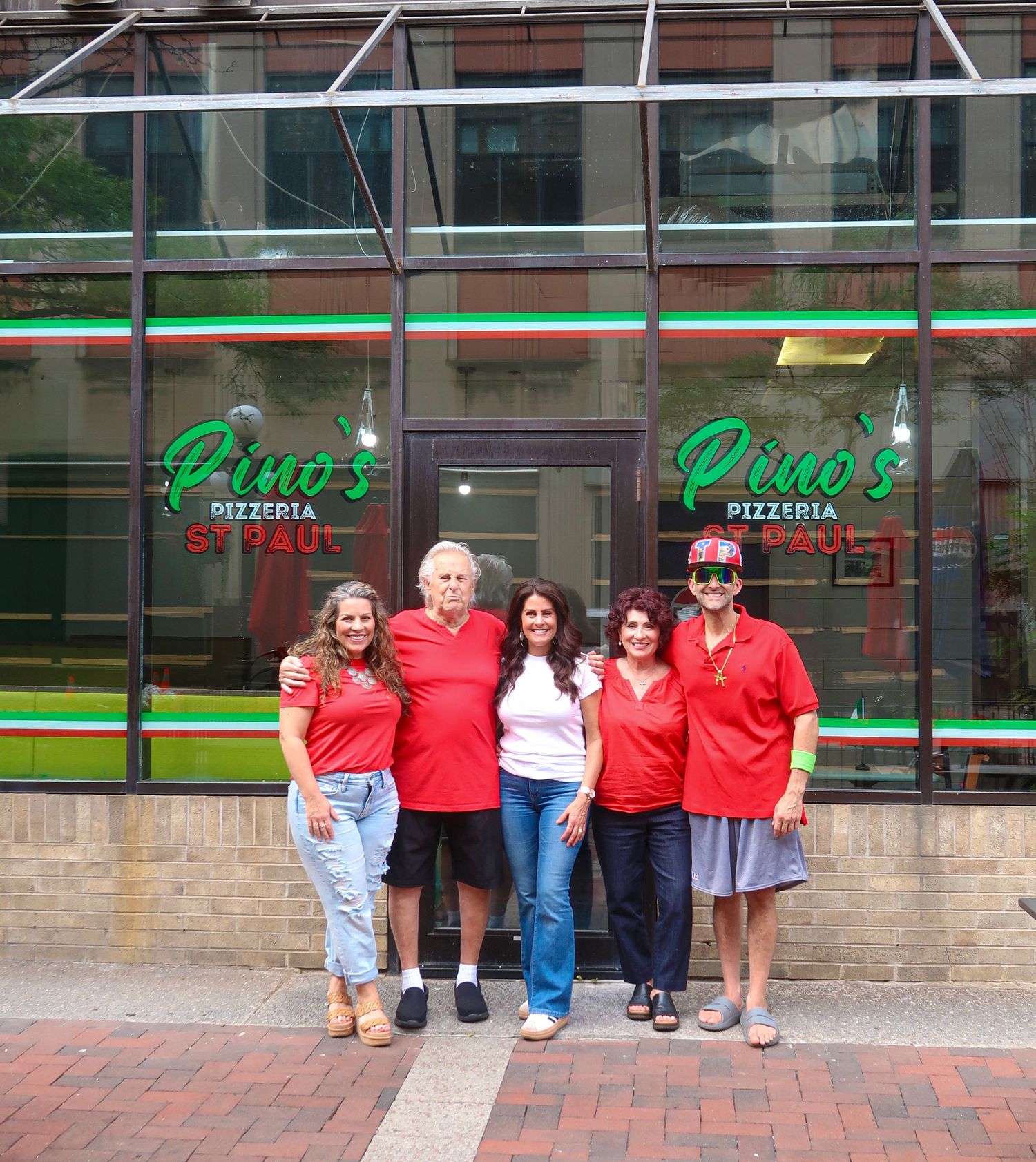 Five people in red shirts pose outside Pino's Pizzeria restaurant entrance on a city sidewalk.