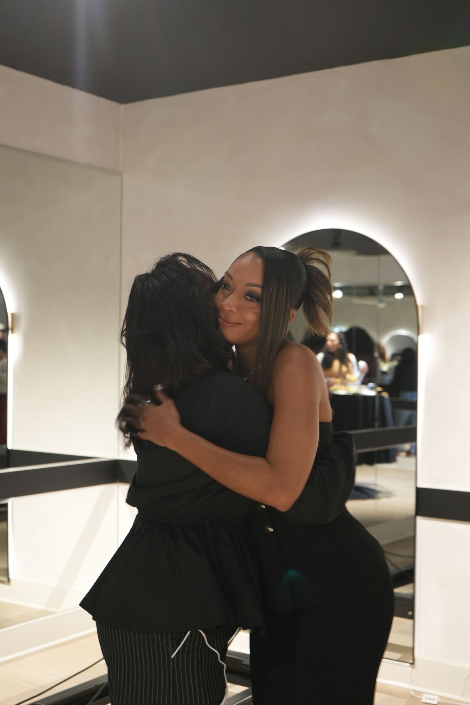 Two women in black outfits sharing a warm hug in a modern salon with arched mirrors and soft lighting.