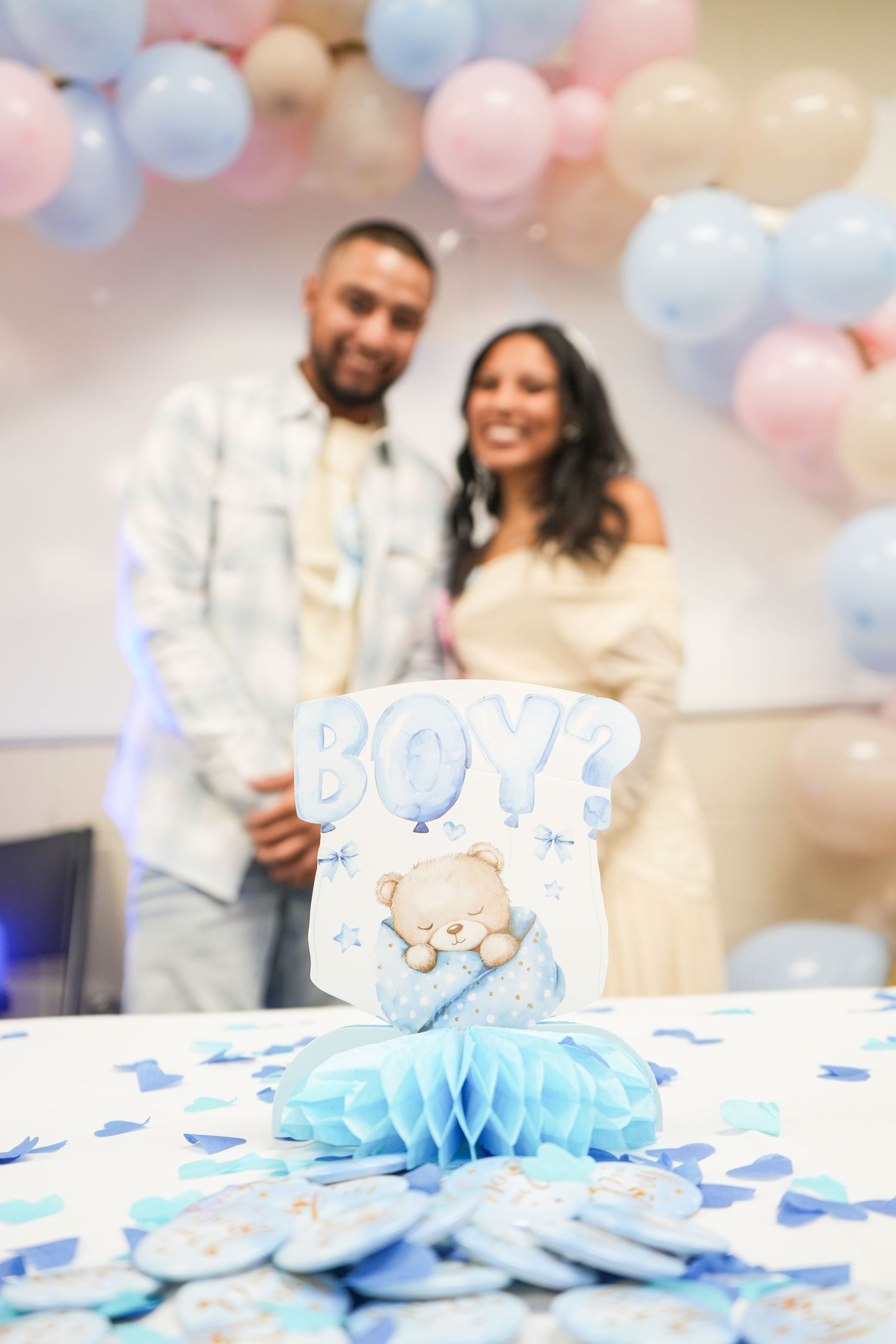 Couple smiling at a gender reveal party with a blue BOY decorative sign, teddy bear, and pink and blue balloons.