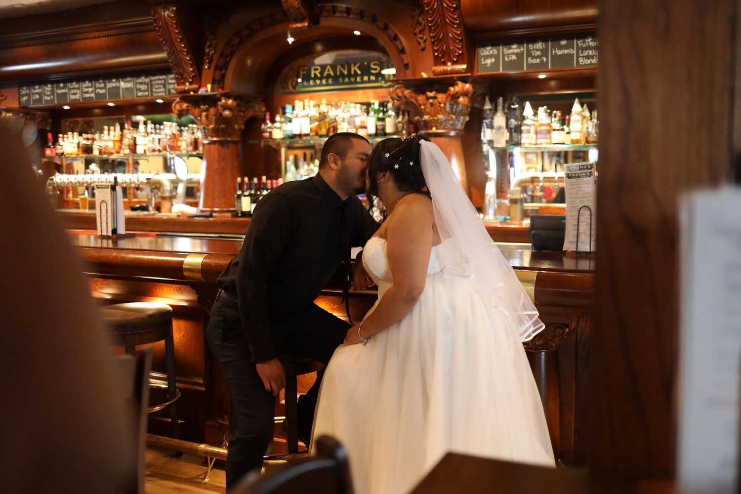 Newlywed couple sharing a romantic moment at a vintage wooden bar, bride in white gown and veil.