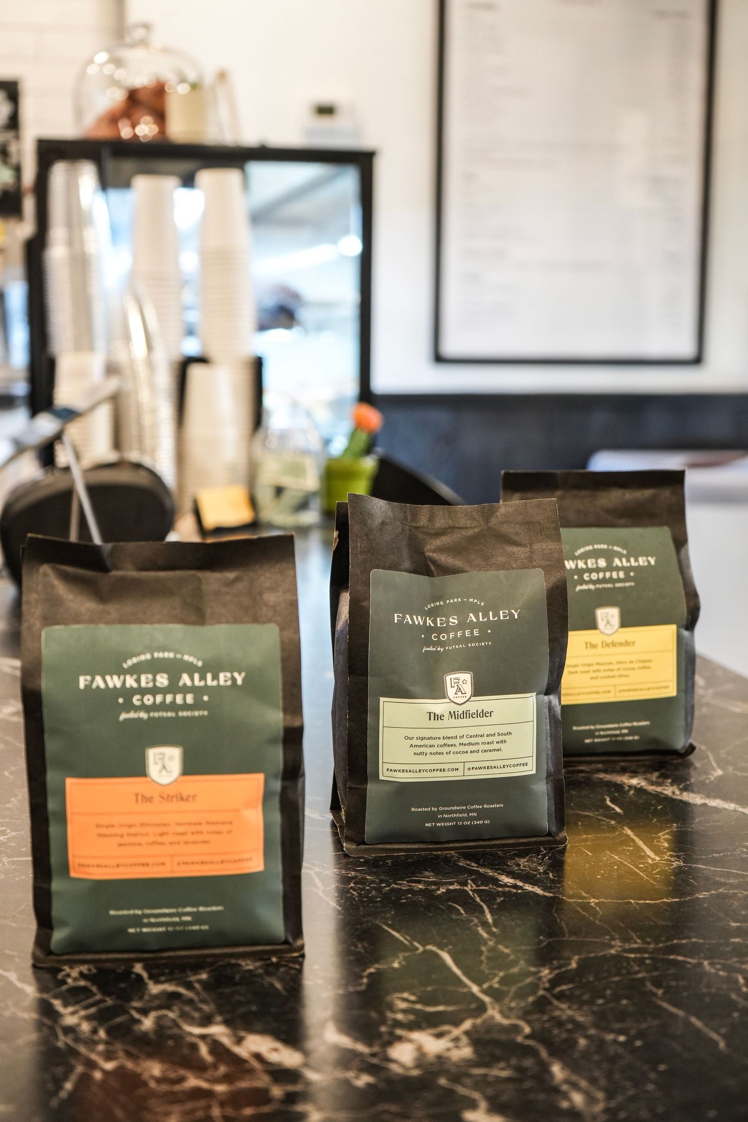 Three bags of Fawkes Alley Coffee on a marble counter in a modern cafe setting.