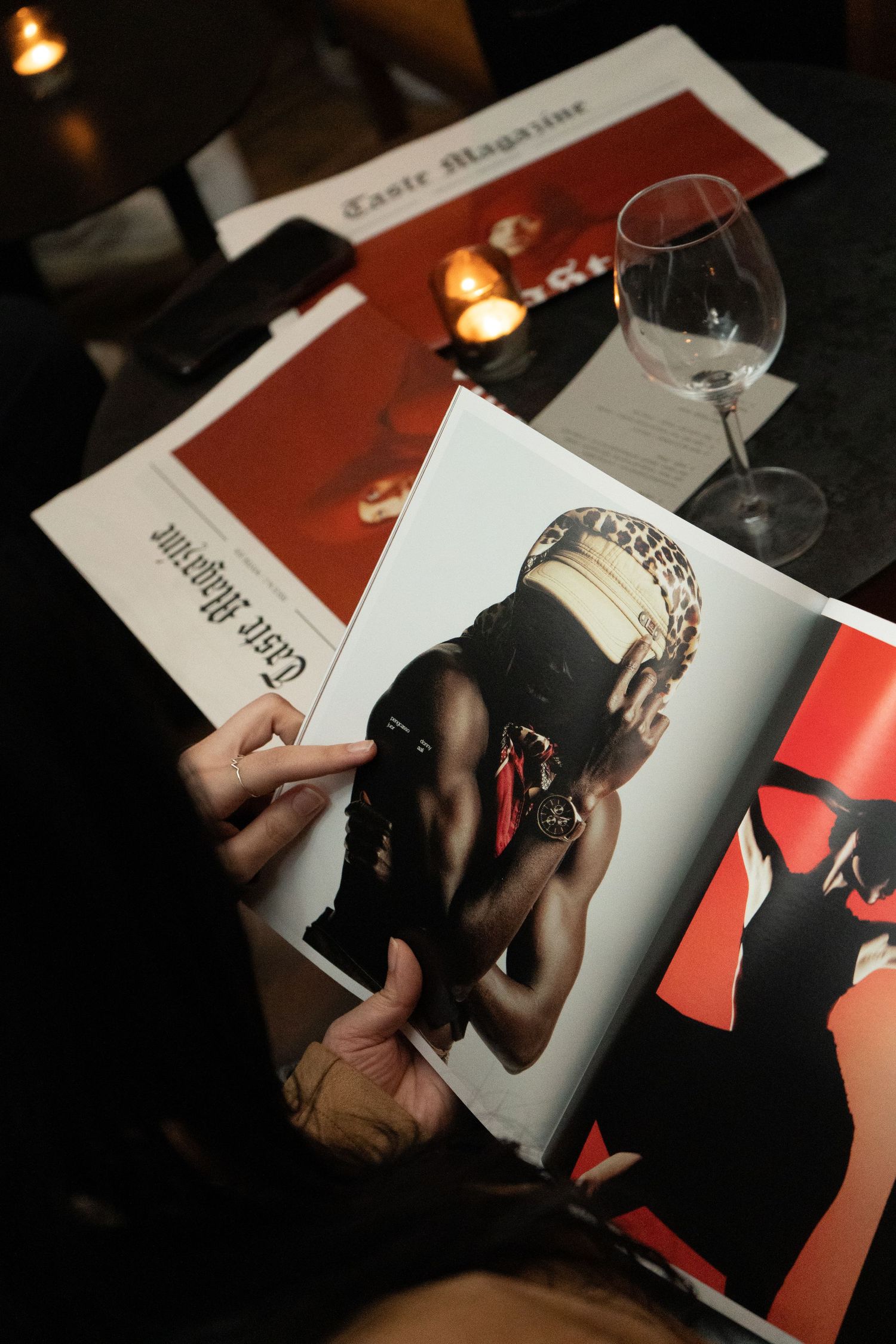 Person holding Taste Magazine open to a fashion editorial page, with wine glass and candles on dark table.