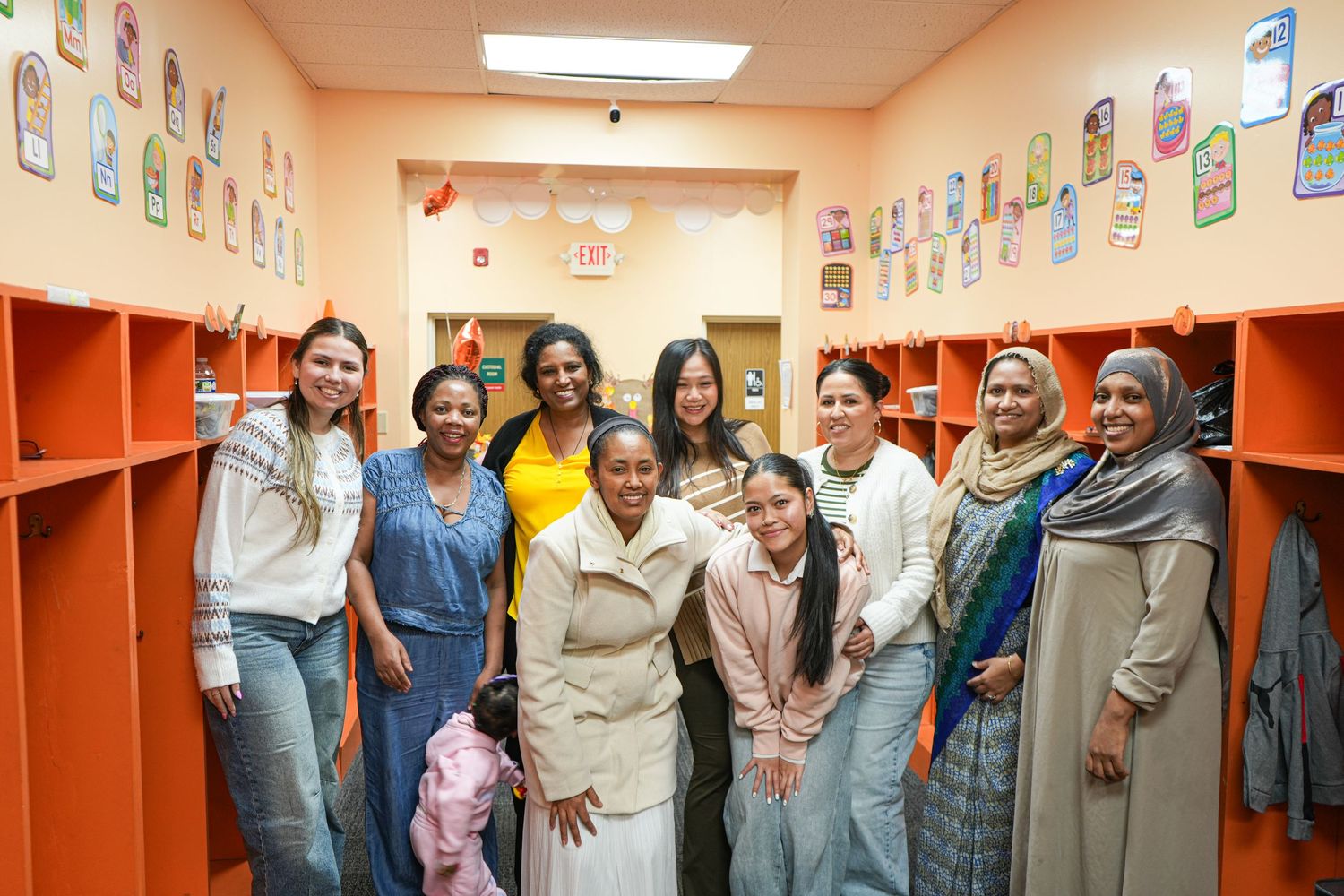 Diverse group of women smiling together in a colorful orange school hallway with lockers and student artwork.