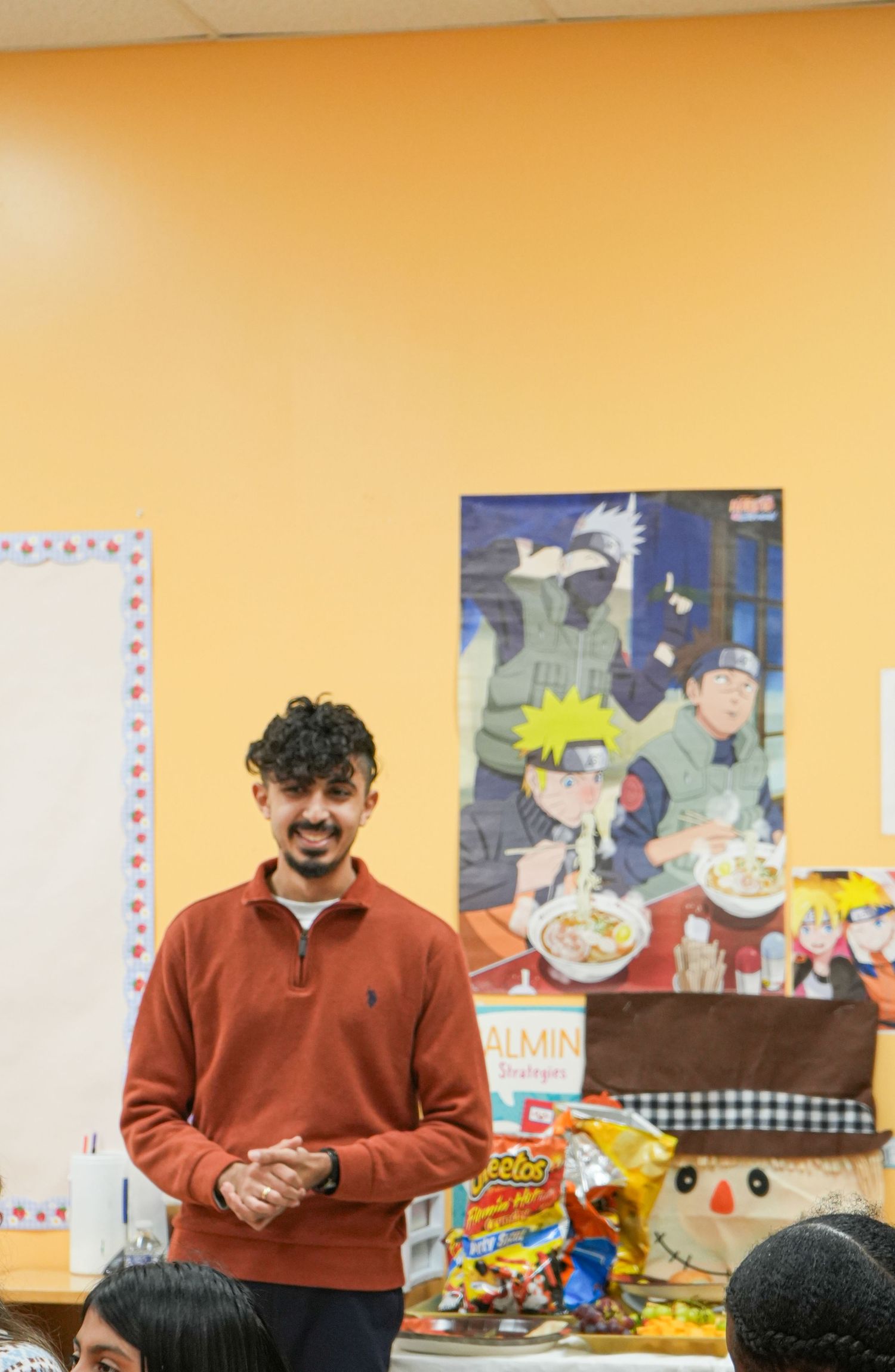 Young man in red sweater smiling in classroom with Naruto anime poster and snacks on table behind him.