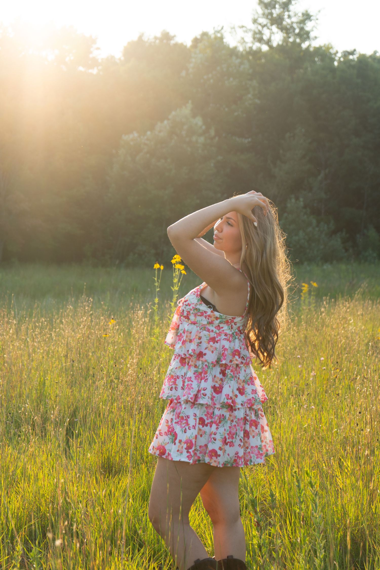 Young woman in floral dress standing in sunlit meadow with wildflowers and trees at golden hour.