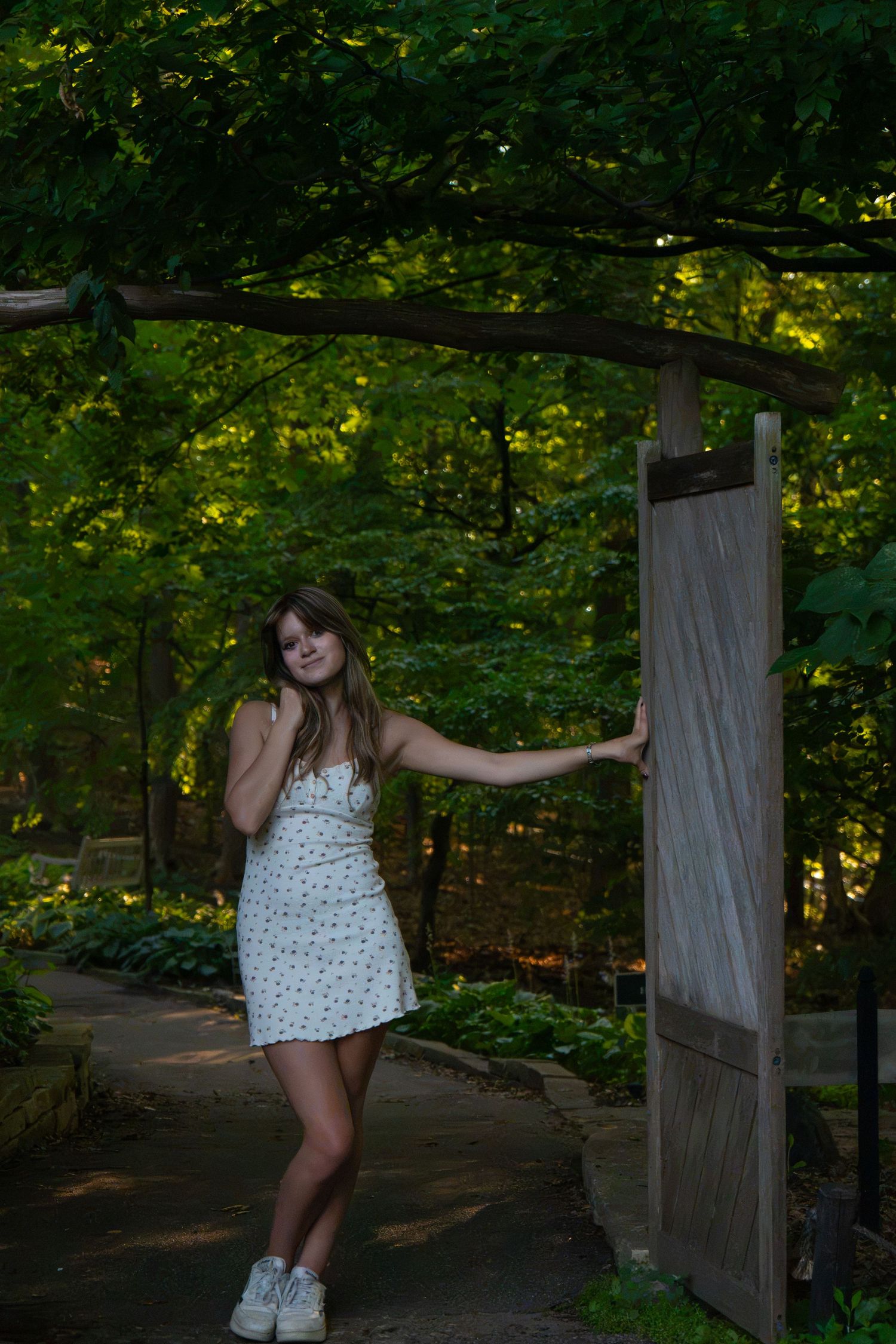 Young woman in white floral sundress posing under rustic wooden garden gate surrounded by lush green forest trees.