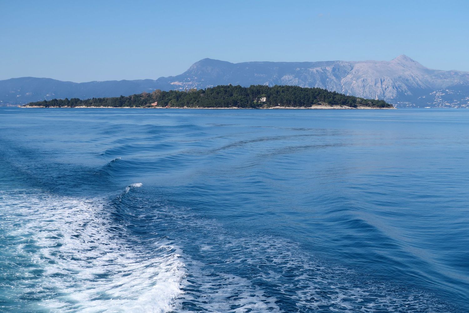 Boat wake trail on calm blue sea with tree-covered island and mountain backdrop under clear sky.