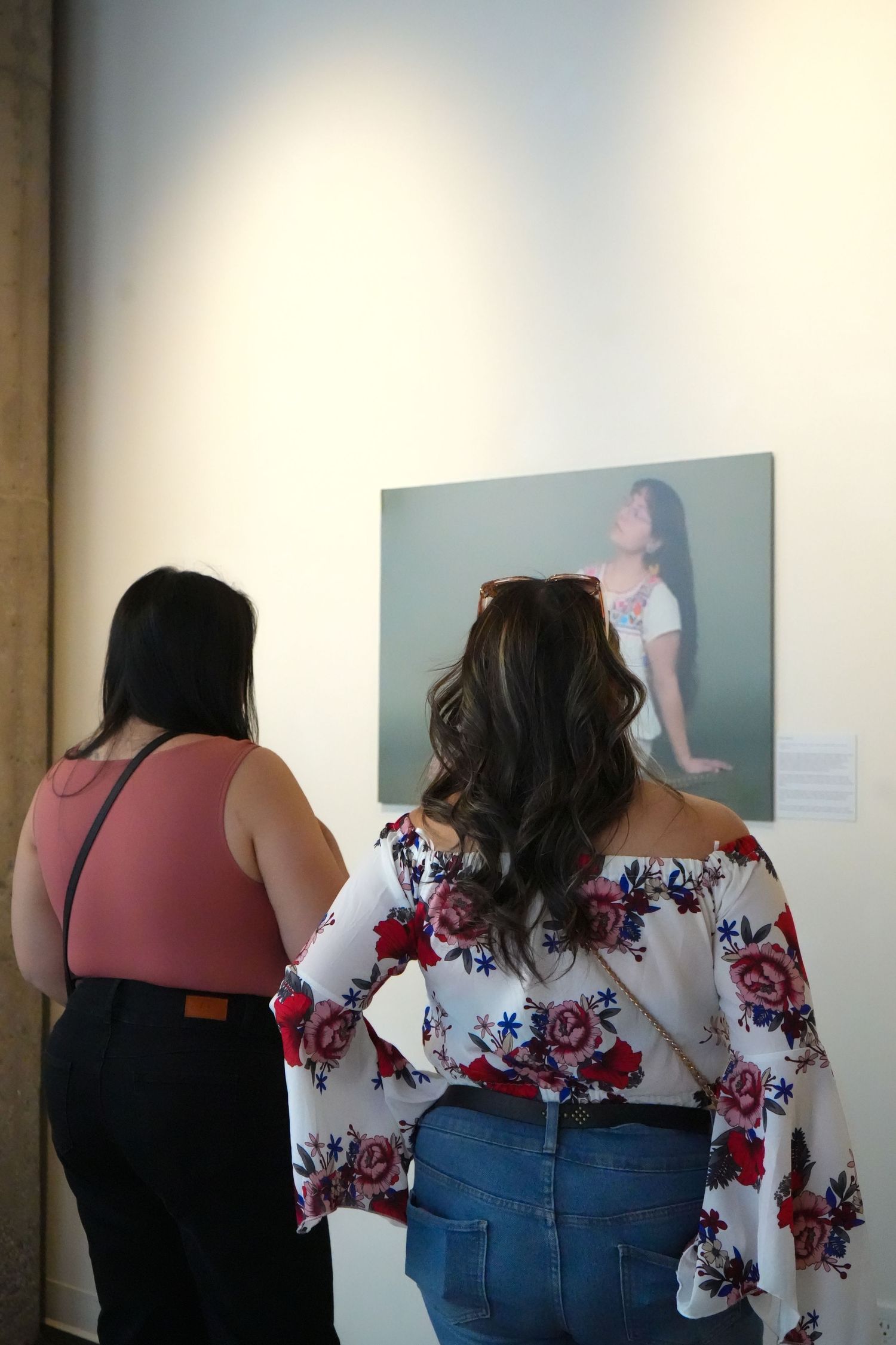 Two women viewing a portrait photograph displayed on a white gallery wall at an art exhibition.