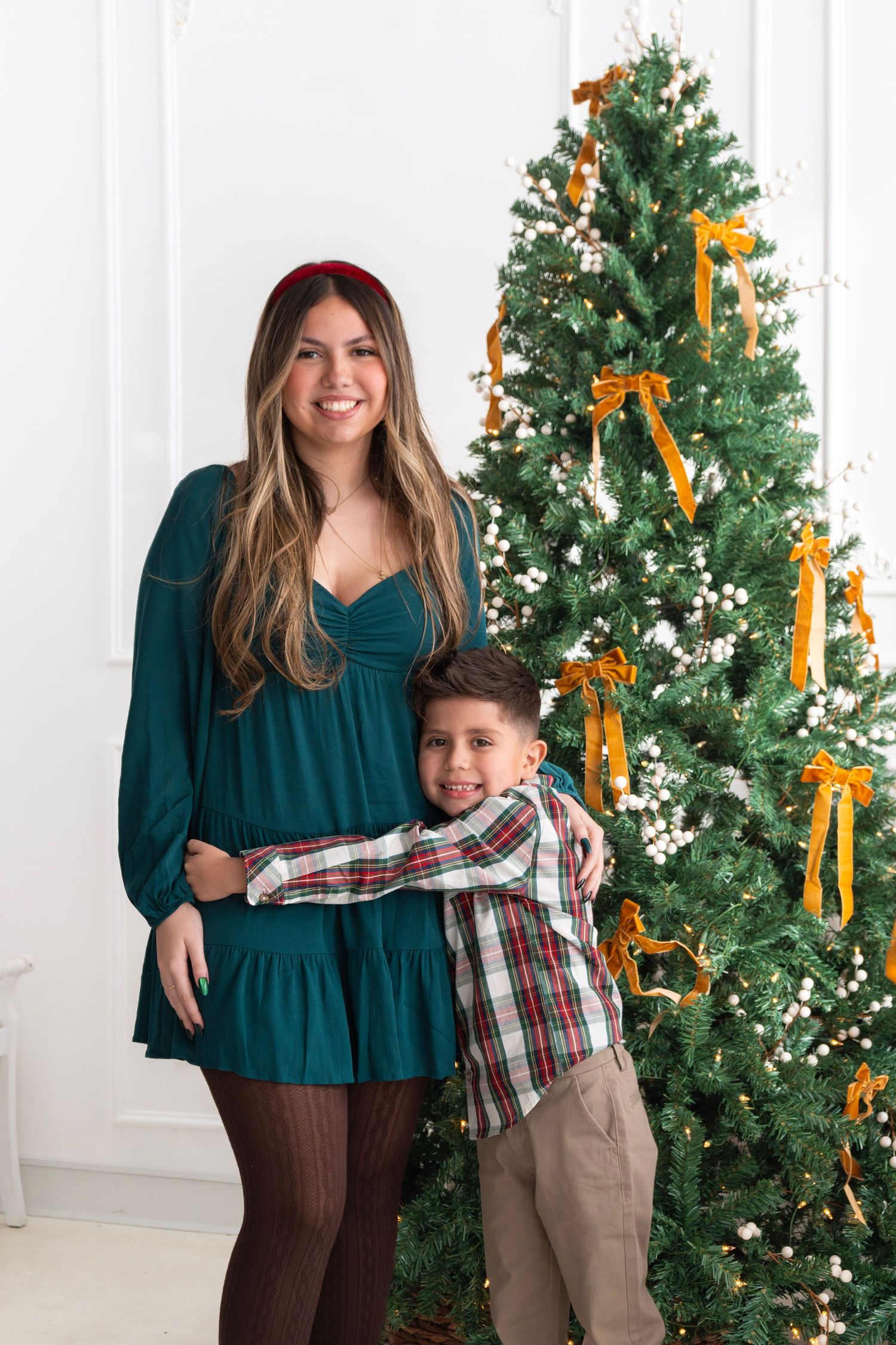 Woman in teal dress and boy in plaid shirt hugging beside decorated Christmas tree with gold bows.