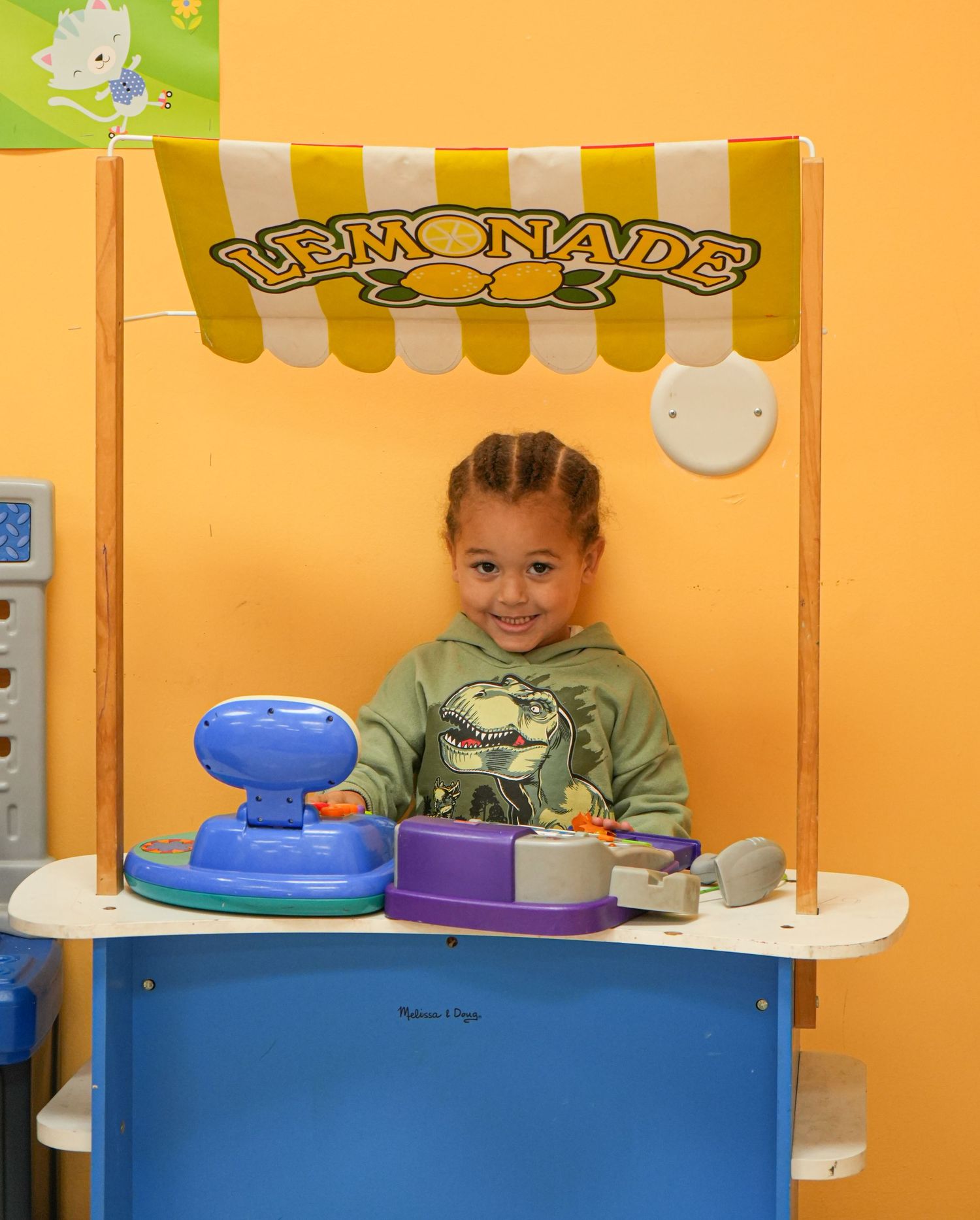Young girl smiling behind a colorful toy lemonade stand with yellow striped awning in a playroom.