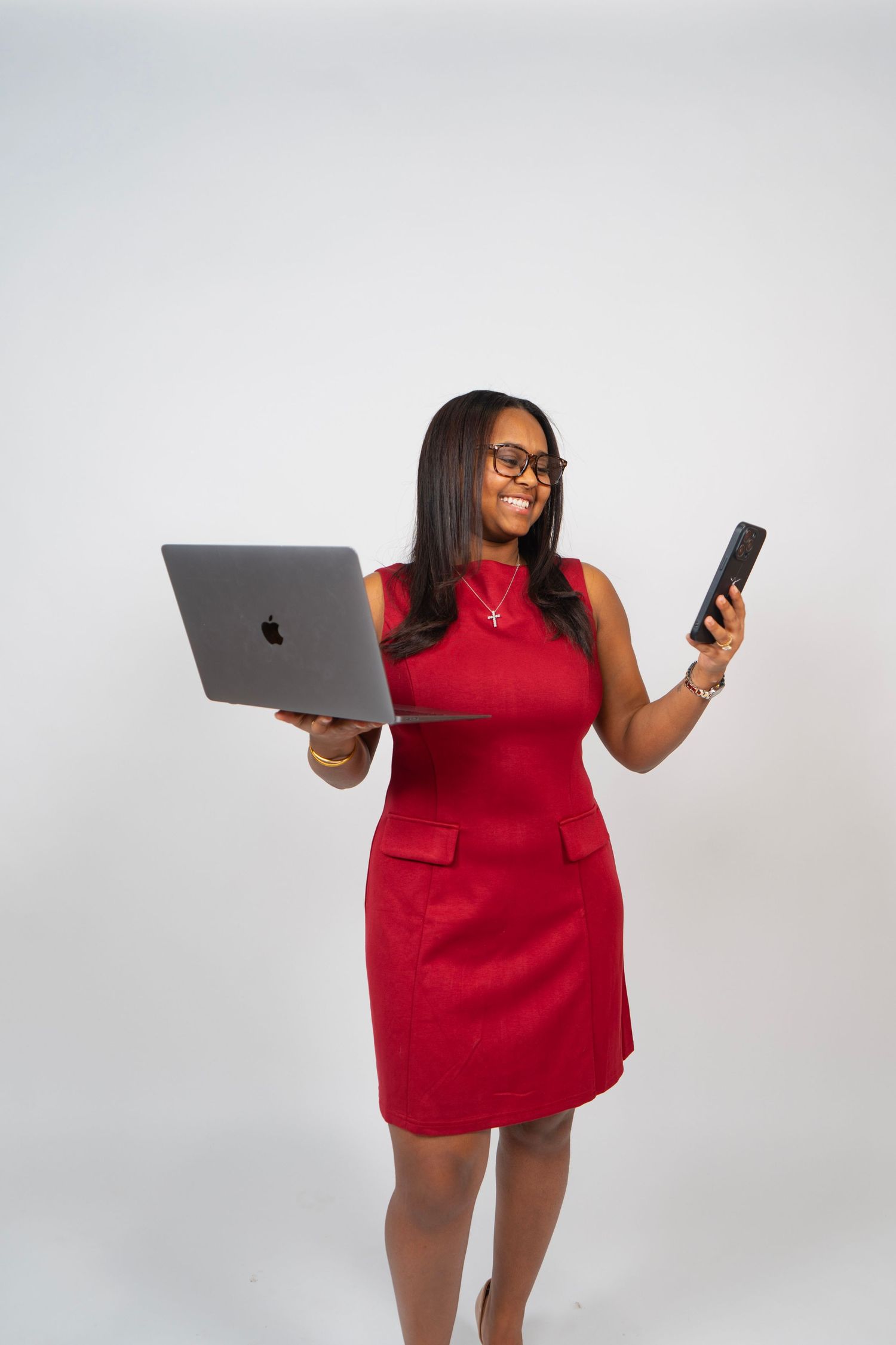 A smiling Black woman in a red dress holds a MacBook laptop and smartphone while posing against a white background.