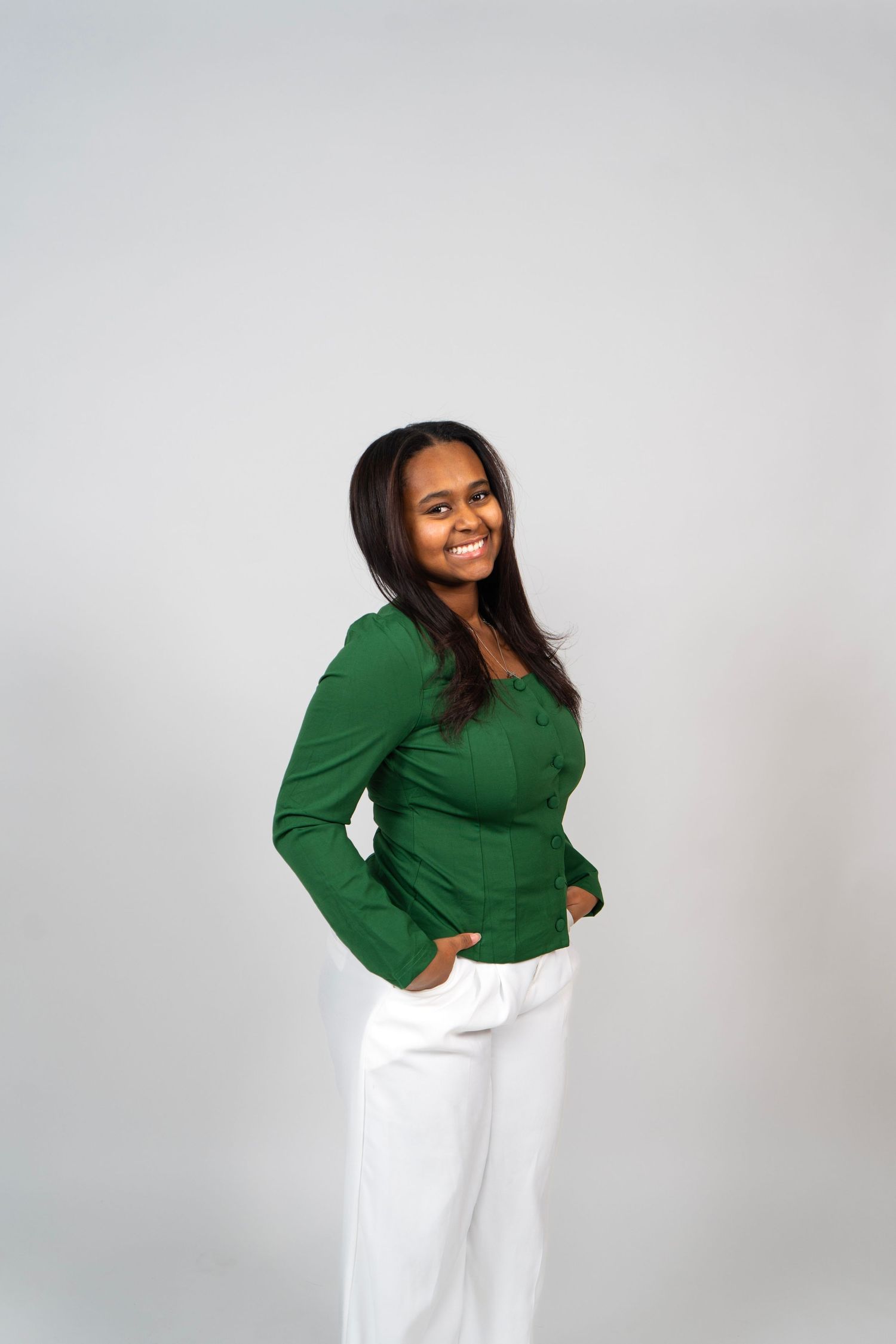 Smiling young woman in green long-sleeve top and white pants posing against a gray background.