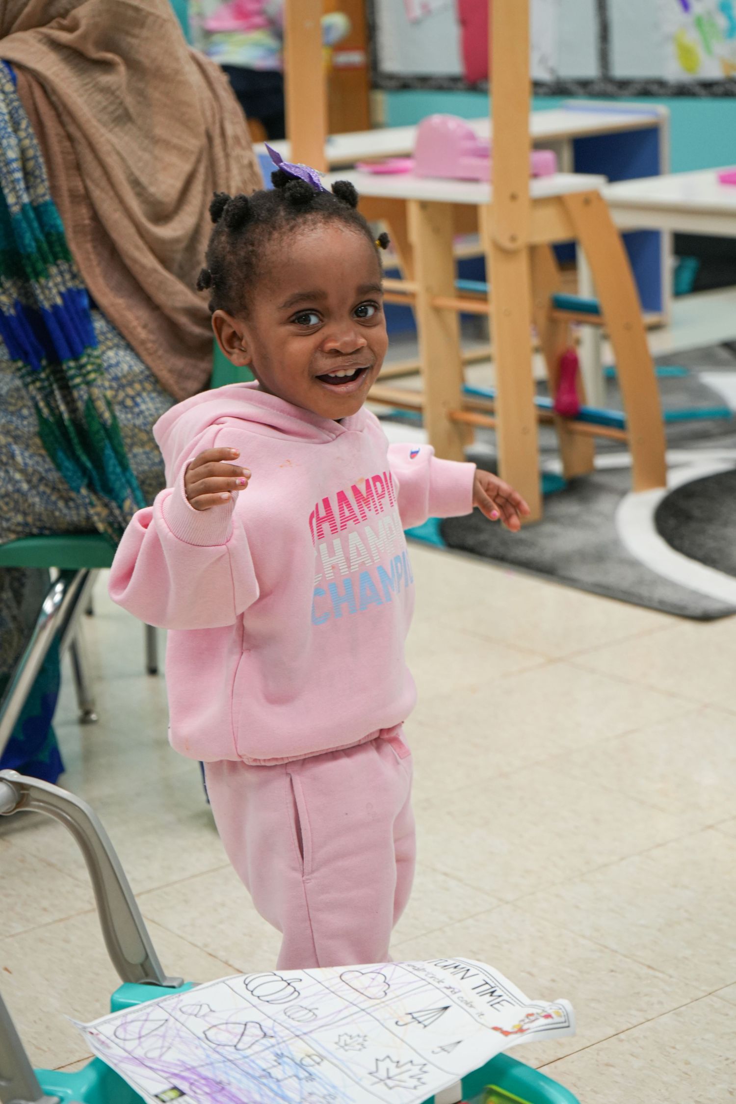 Happy young girl in pink Champion hoodie smiling in a colorful classroom setting.