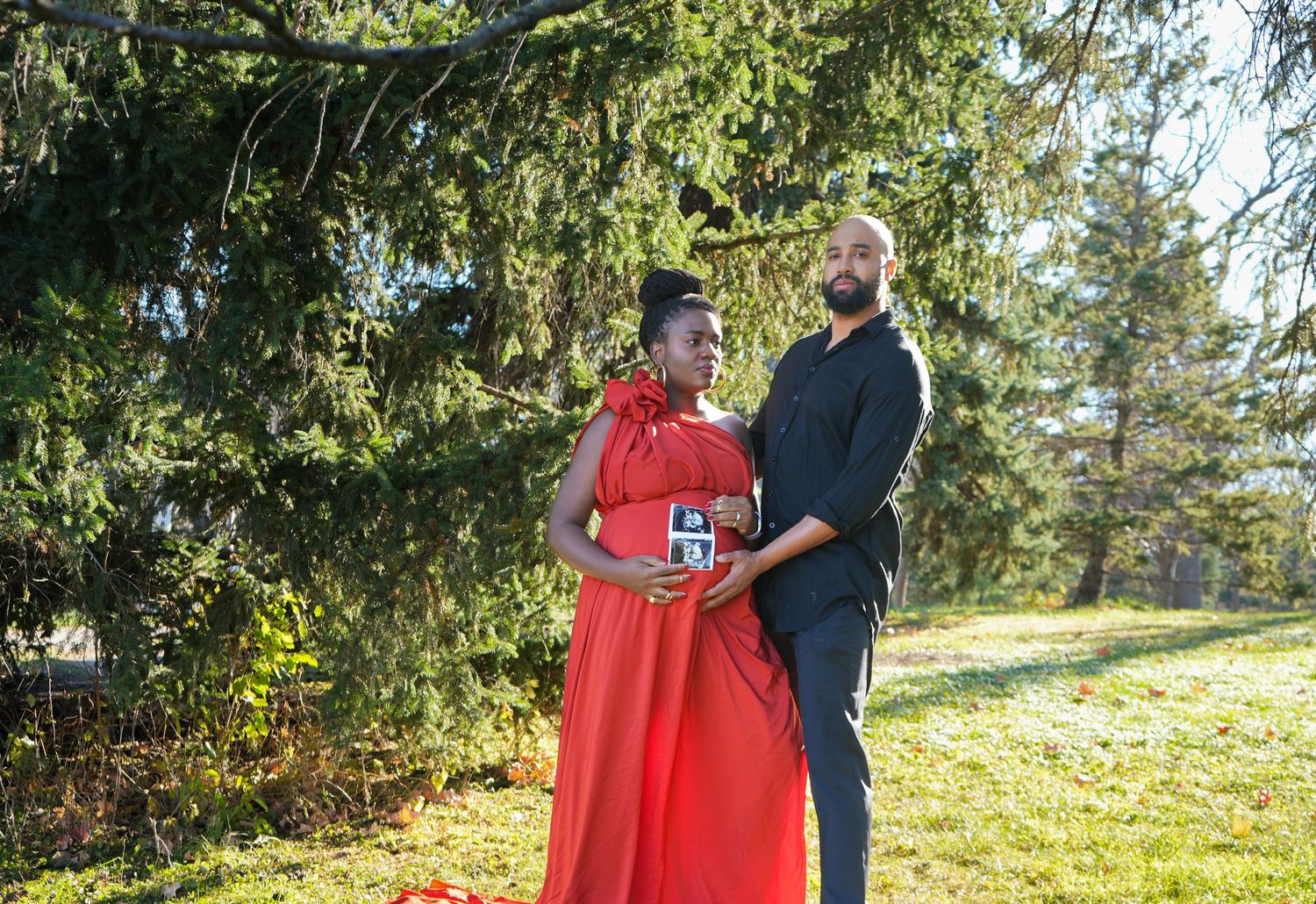 Pregnant woman in red dress and man in black outfit pose together outdoors among green trees on a sunny day.
