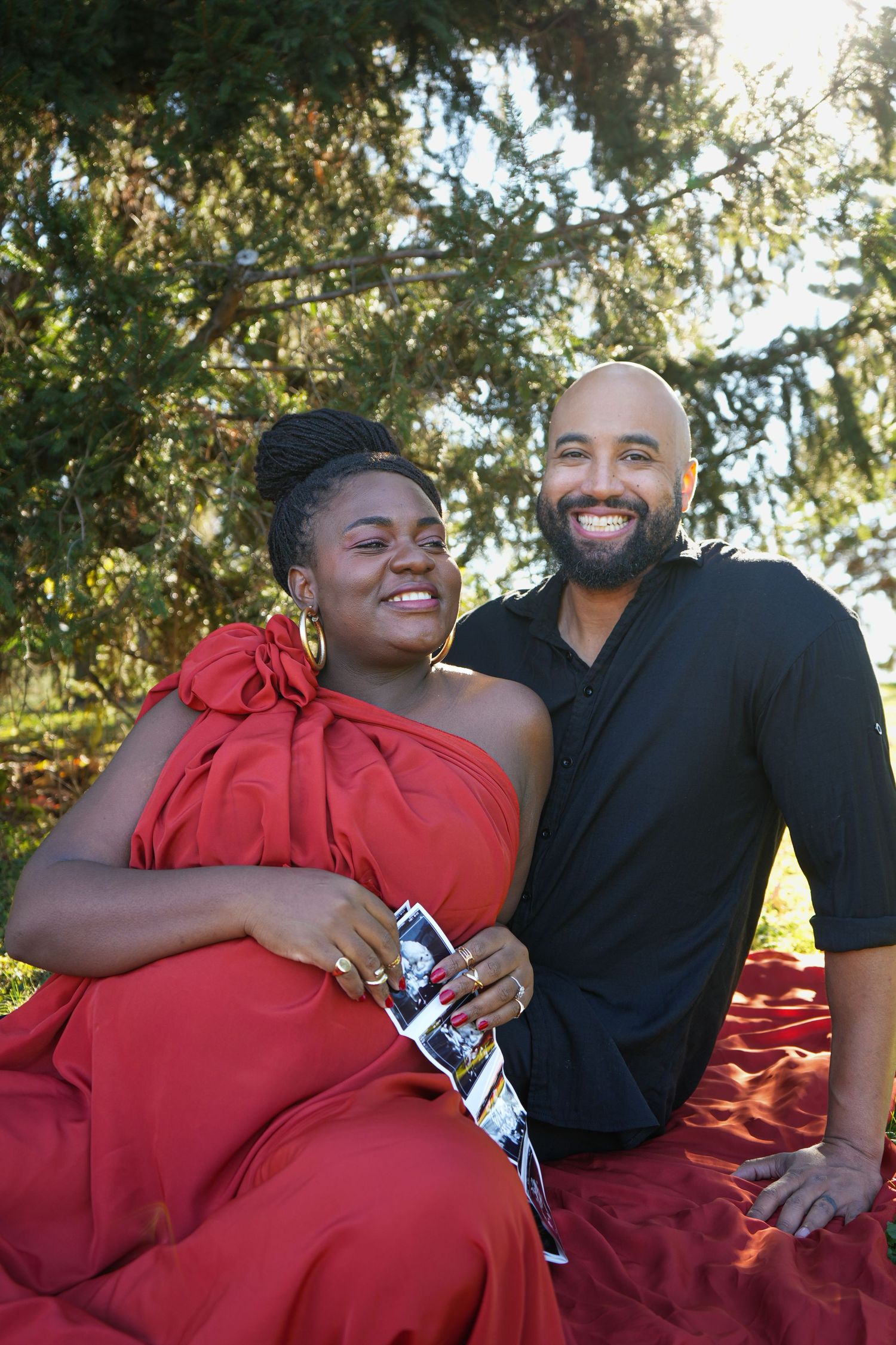 Smiling pregnant woman in red dress holds ultrasound photo with her partner outdoors on a red blanket.
